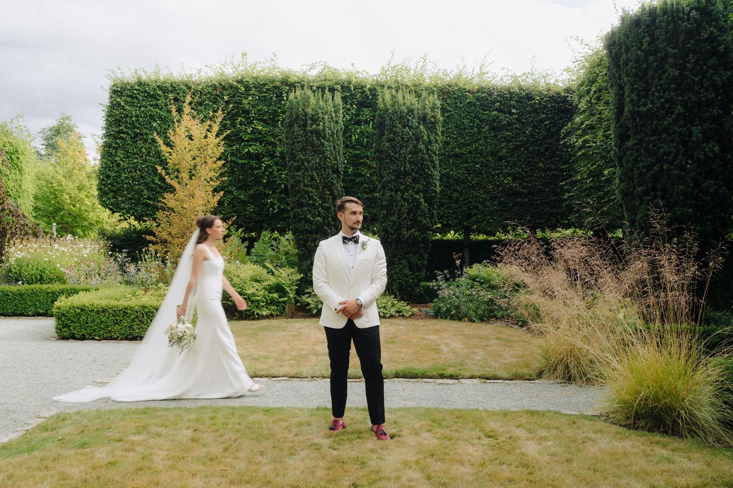 A couple walks along a garden path in elegant wedding attire during an outdoor celebration.