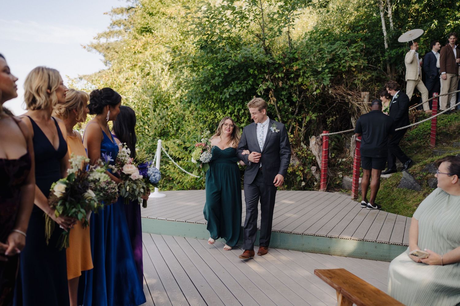 Outdoor wedding ceremony setup with guests seated on wooden deck surrounded by lush greenery.