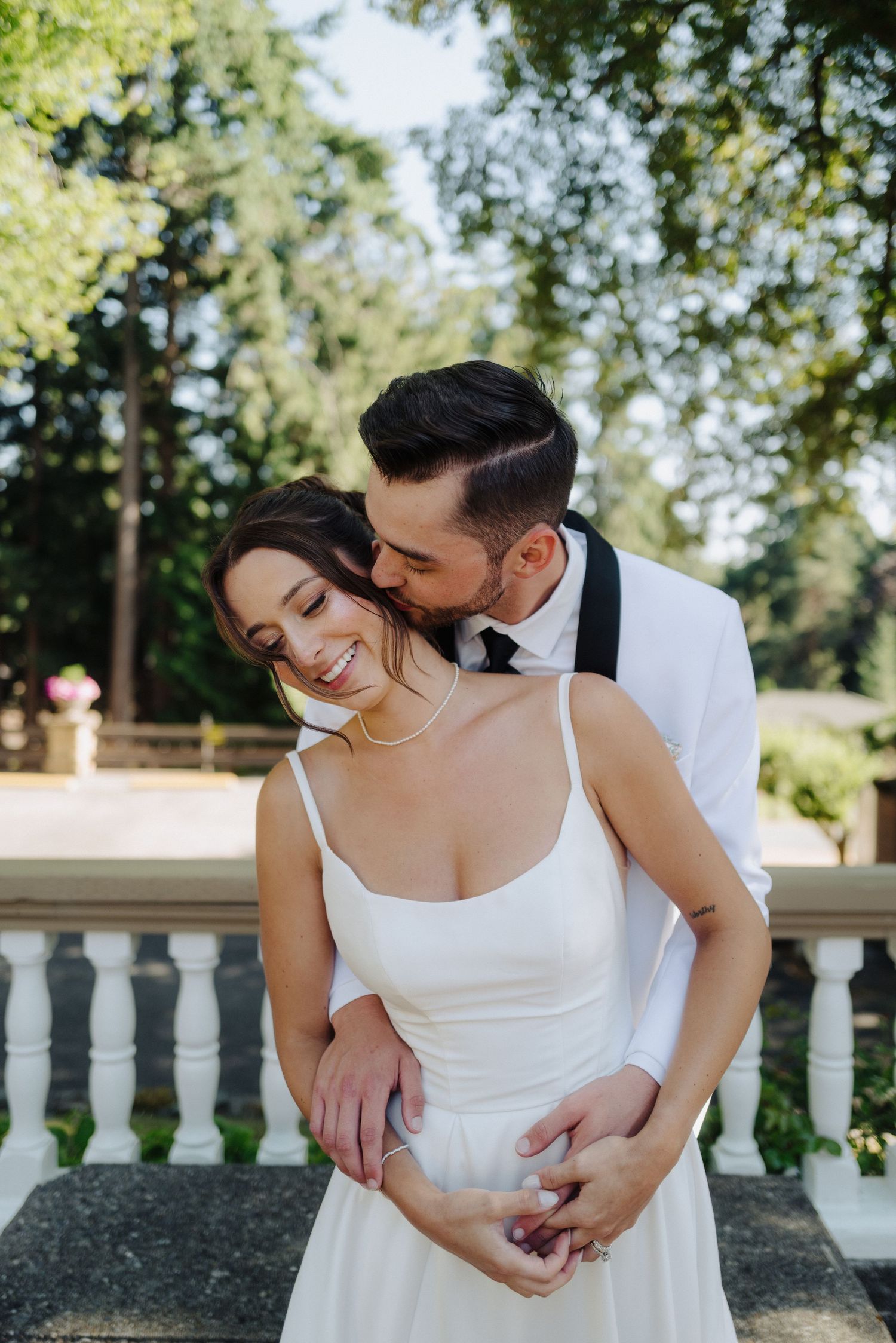 A romantic couple in wedding attire embraces on a sunny day by a white stone balustrade in a garden setting.