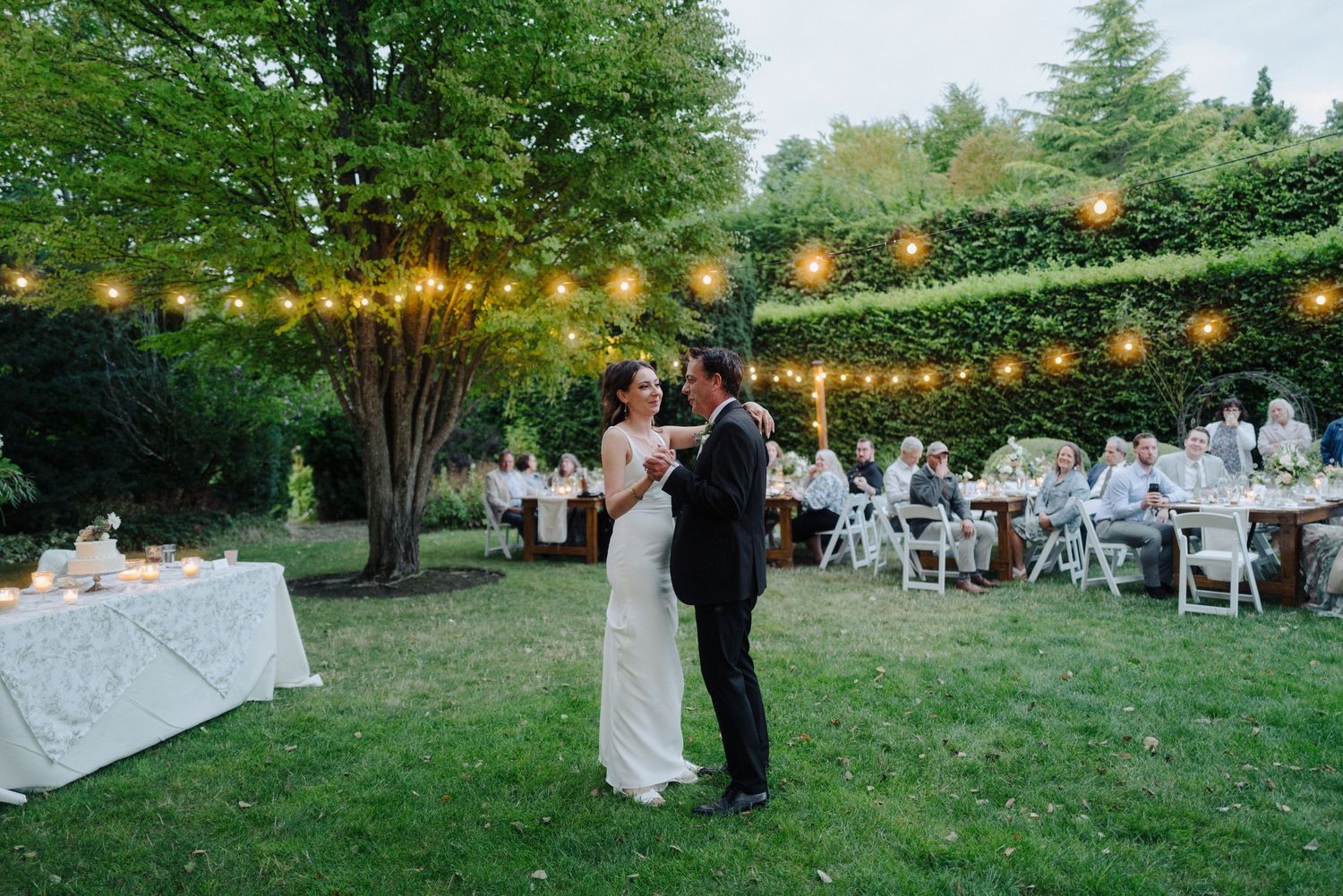 Outdoor wedding reception setup with string lights and white tables under green trees.