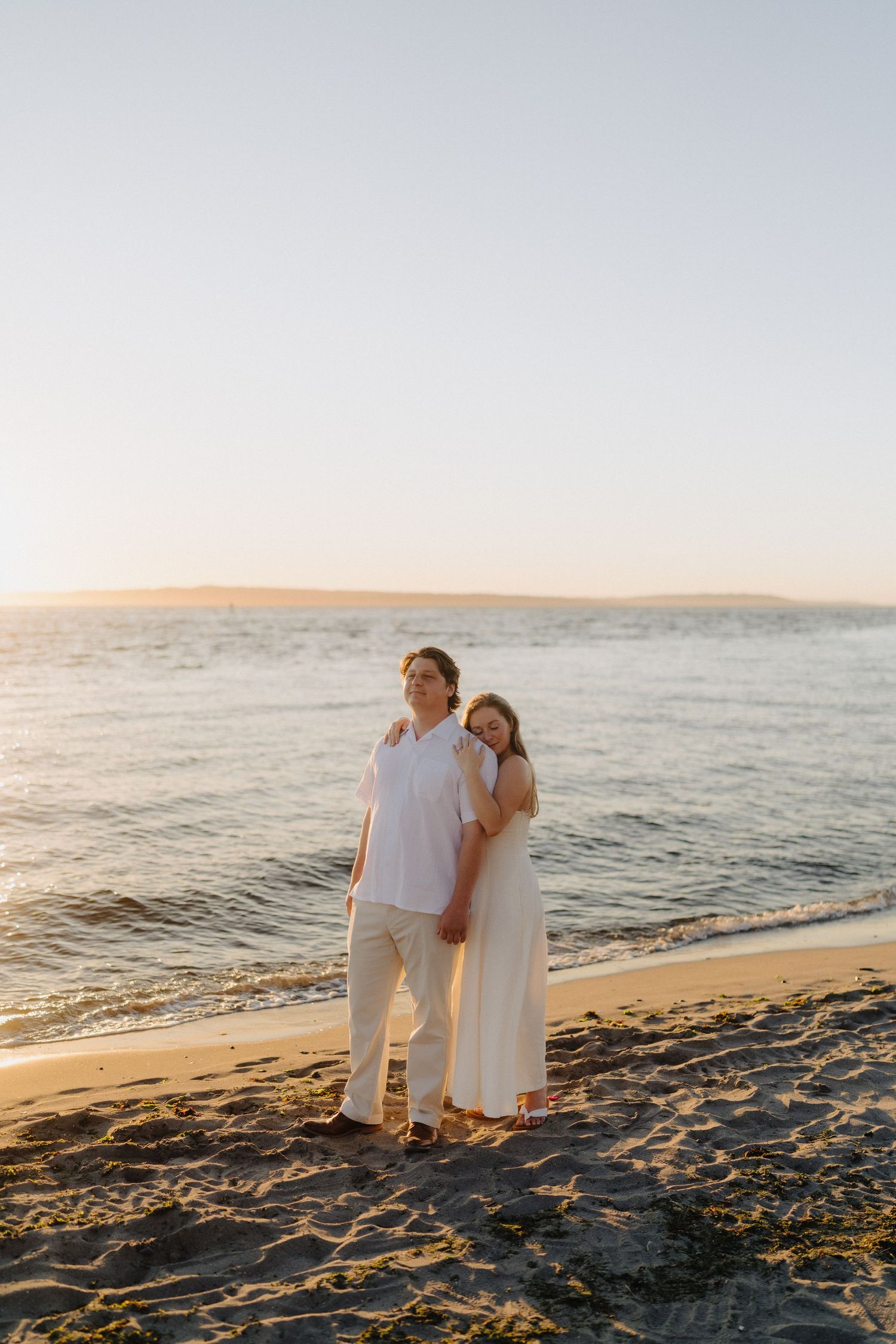A couple in white outfits stands together on a beach at sunset, gazing at the ocean waves.