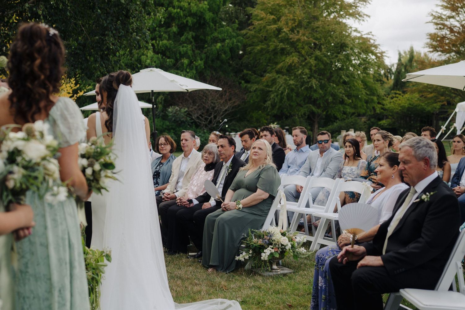 Wedding ceremony guests seated in white chairs on a lawn with umbrellas overhead.