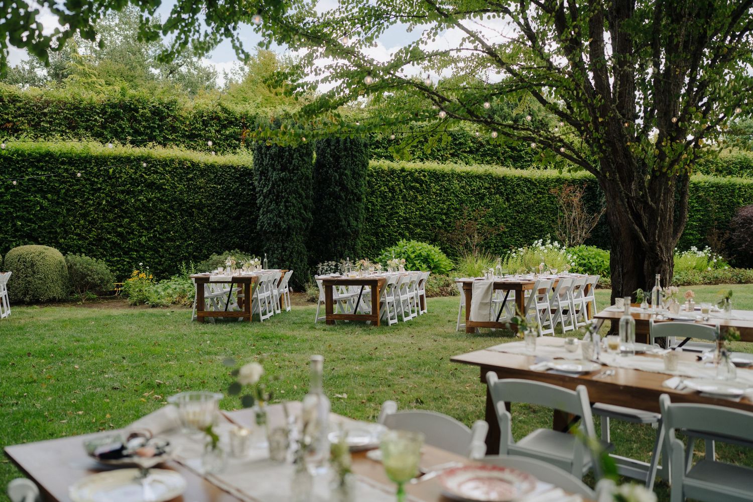 Outdoor wedding reception setup with wooden tables and chairs against manicured garden hedges.