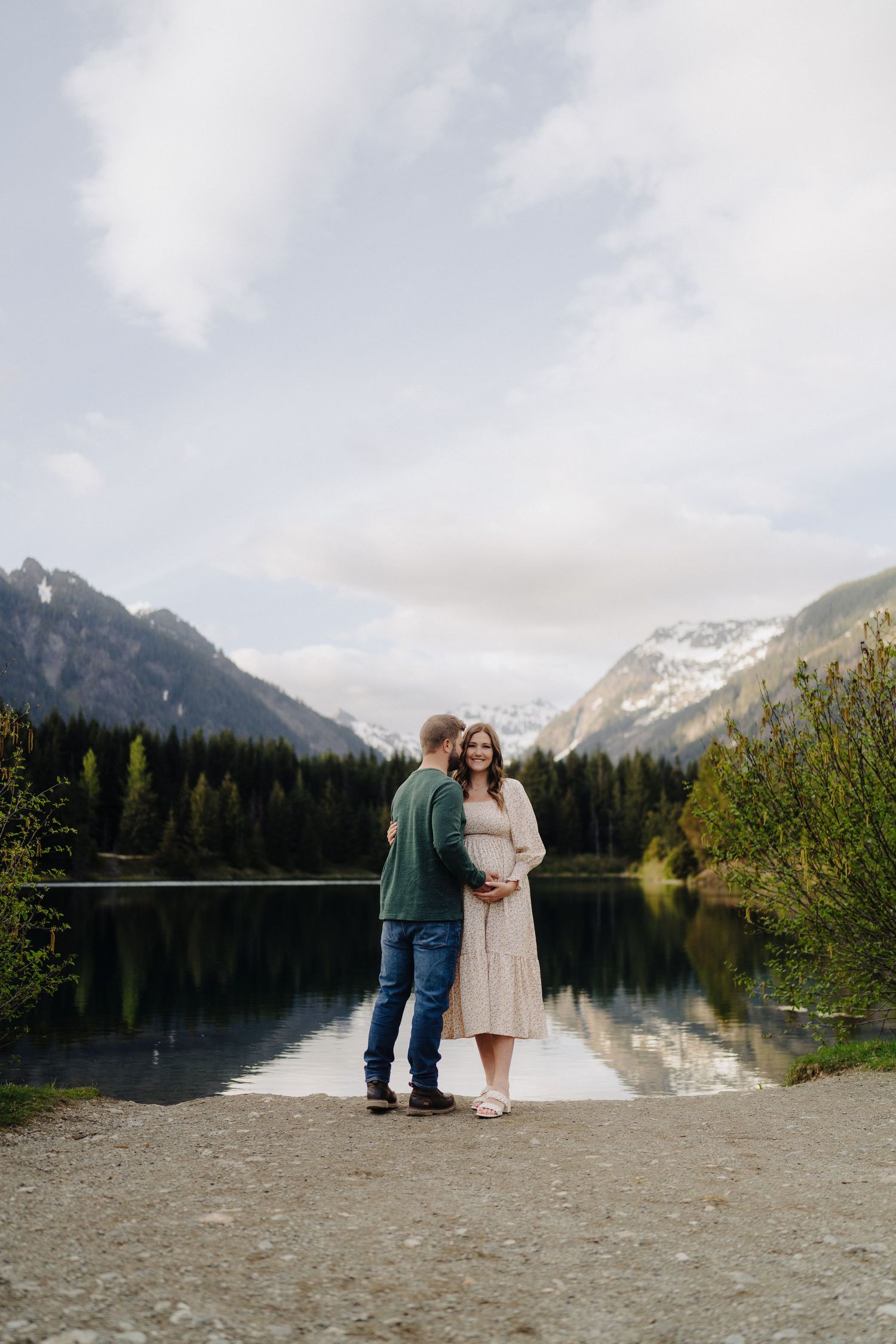 A couple stands by a serene mountain lake surrounded by evergreen forest and snow-capped peaks at sunset.