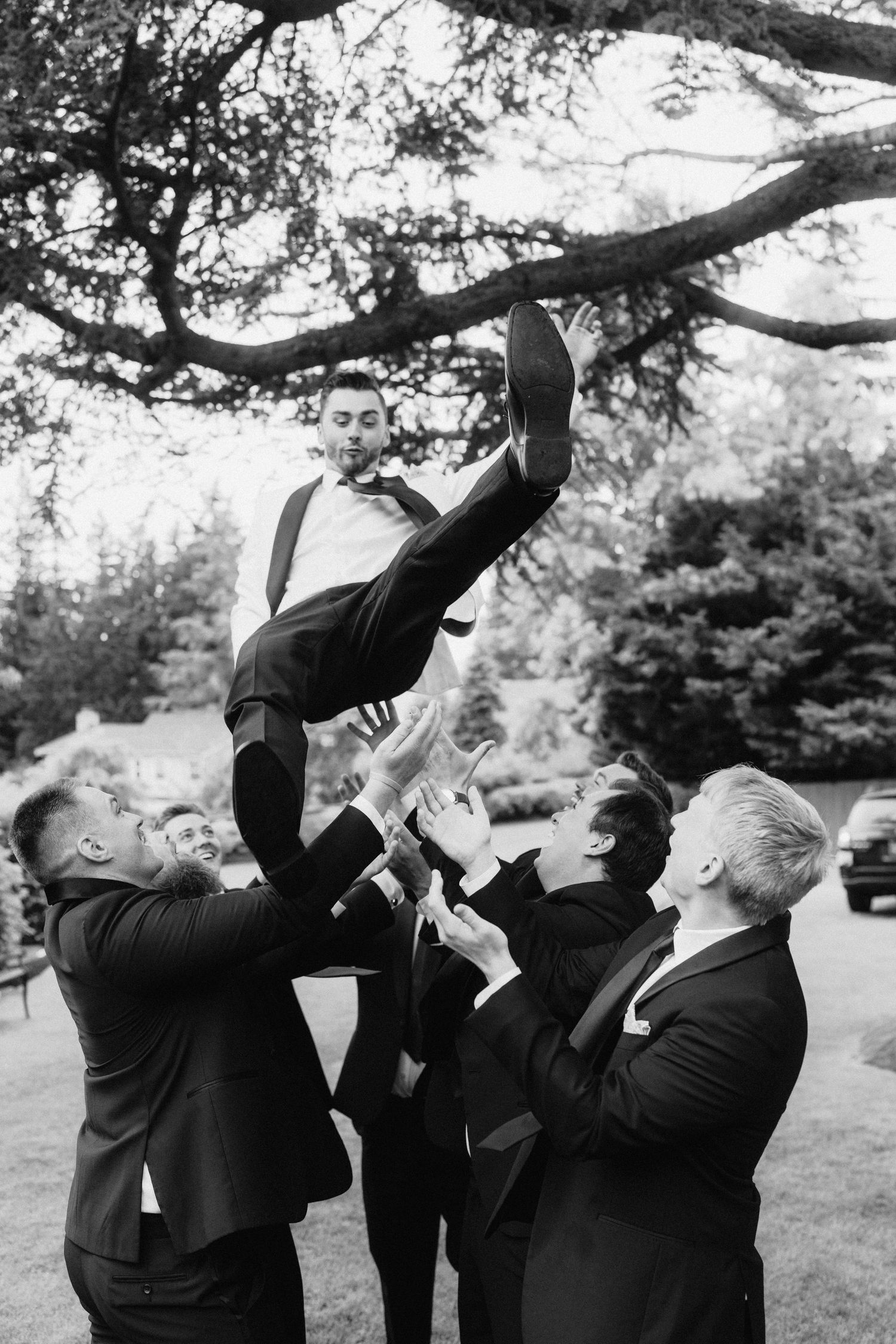 Groomsmen in black suits drinking together outdoors in a synchronized line against trees.
