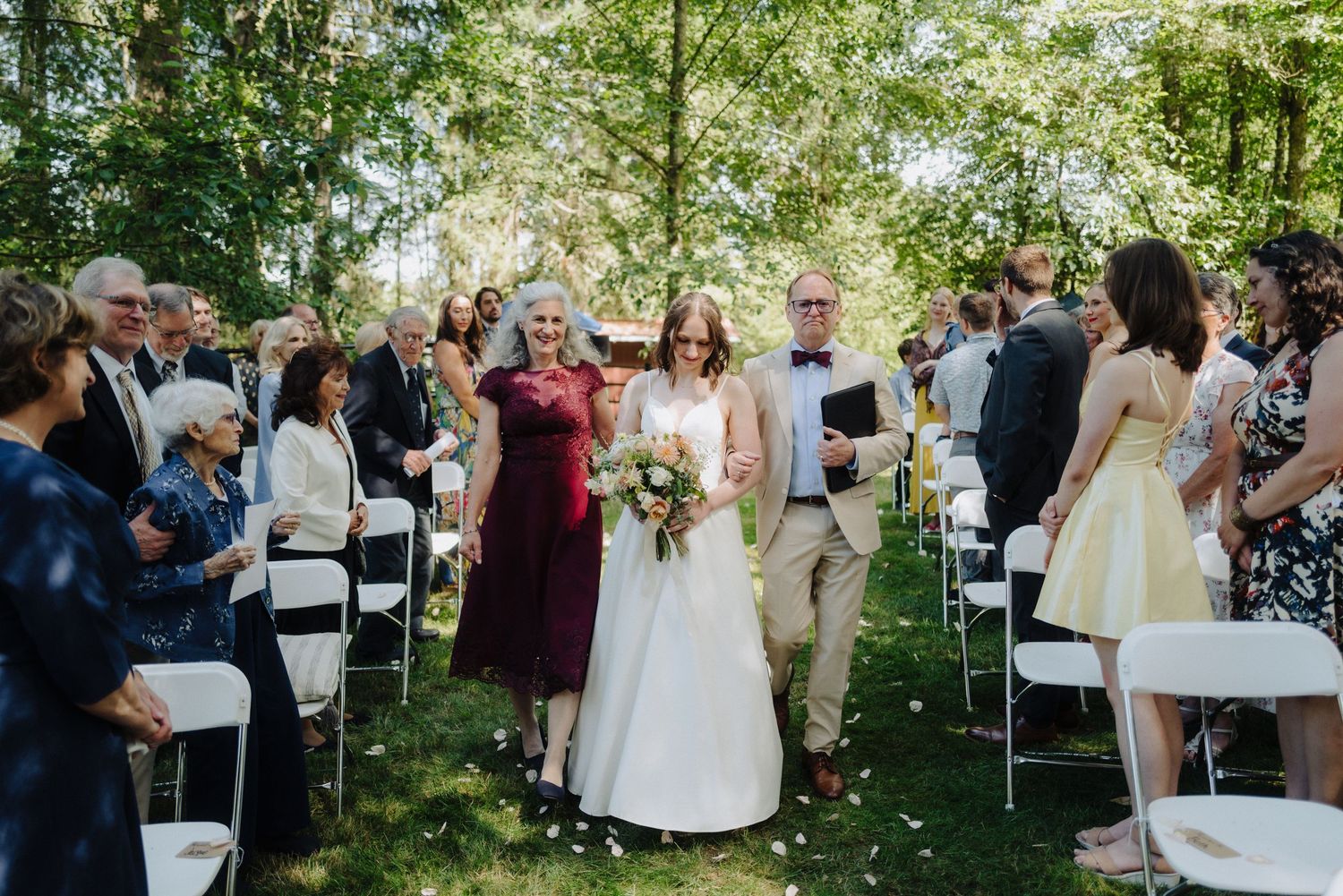 Wide angle view of outdoor wedding ceremony with guests seated among trees and greenery.