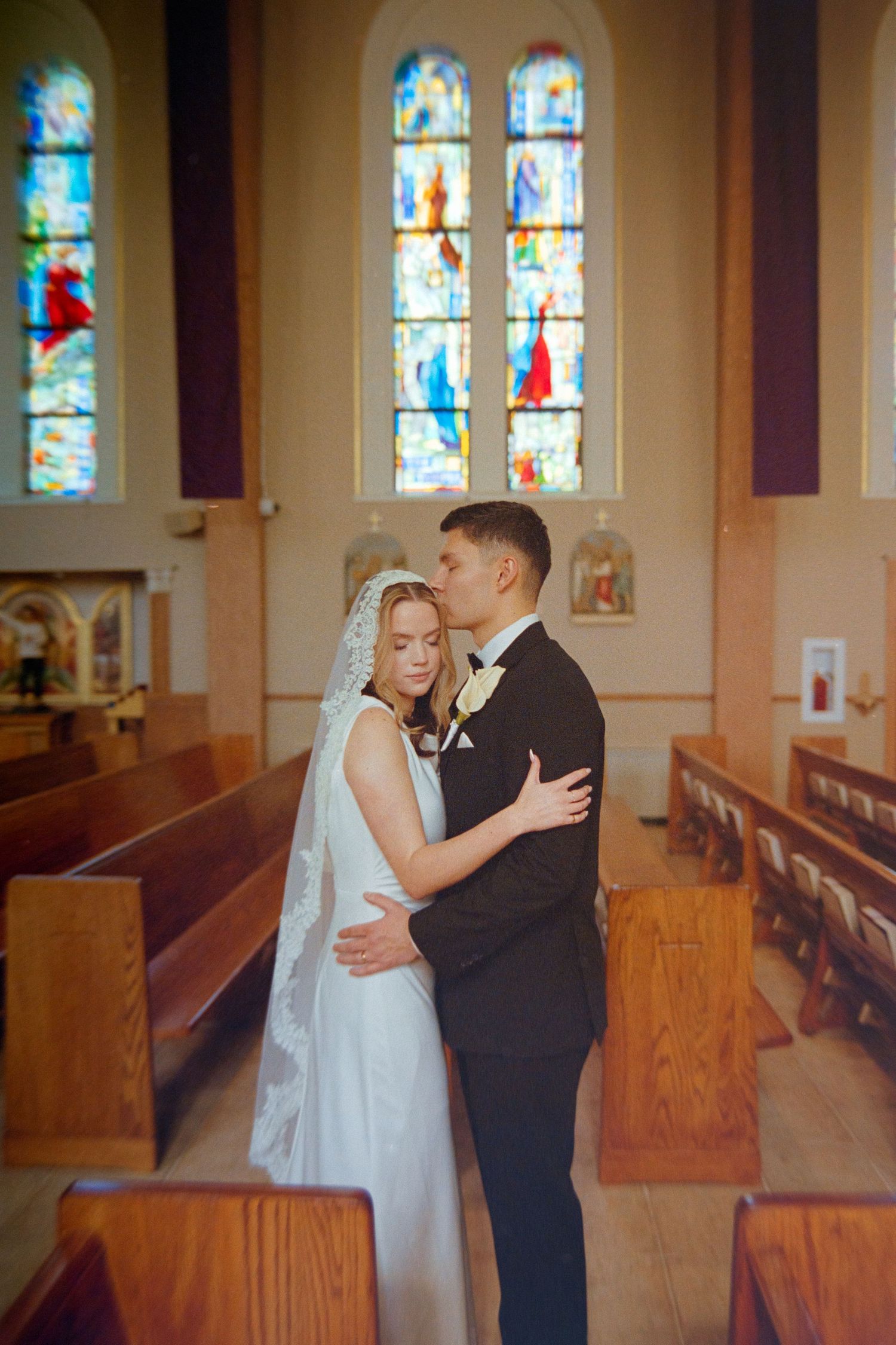 Bride and groom embrace tenderly inside a church with colorful stained glass windows.