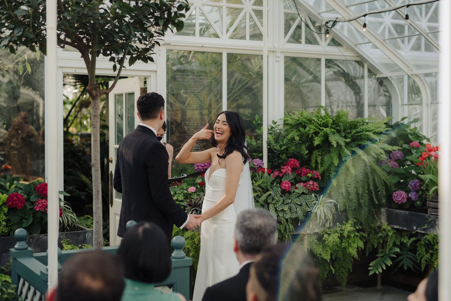 A wedding ceremony takes place inside a greenhouse filled with lush plants and flowers.