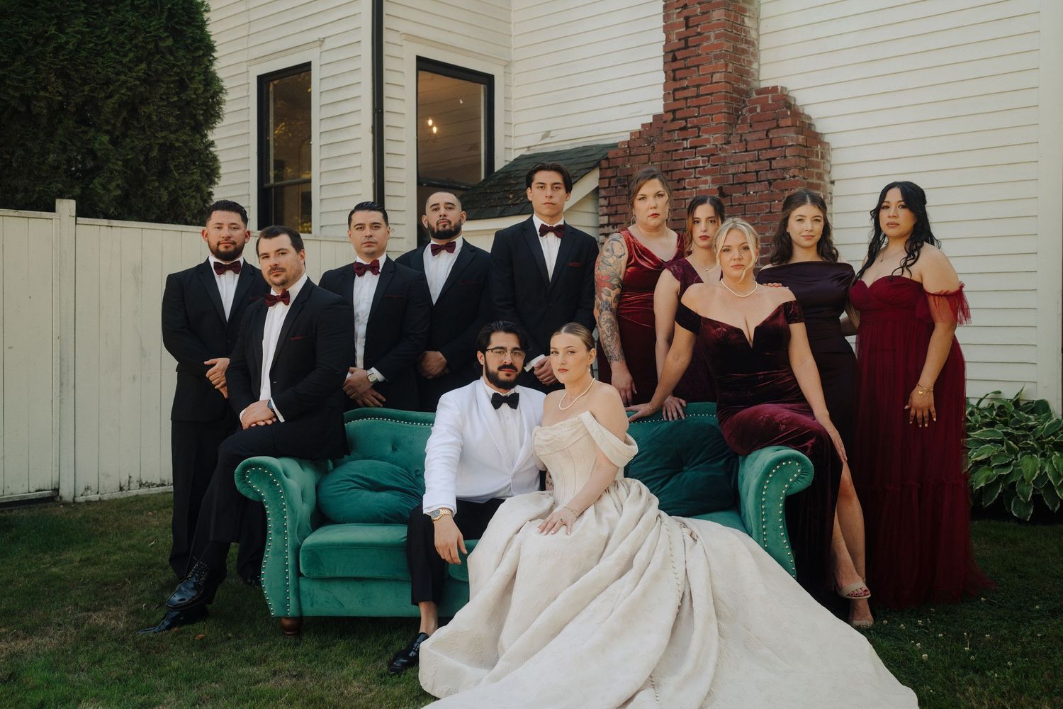 Wedding party poses together on emerald green velvet couch against white house exterior.
