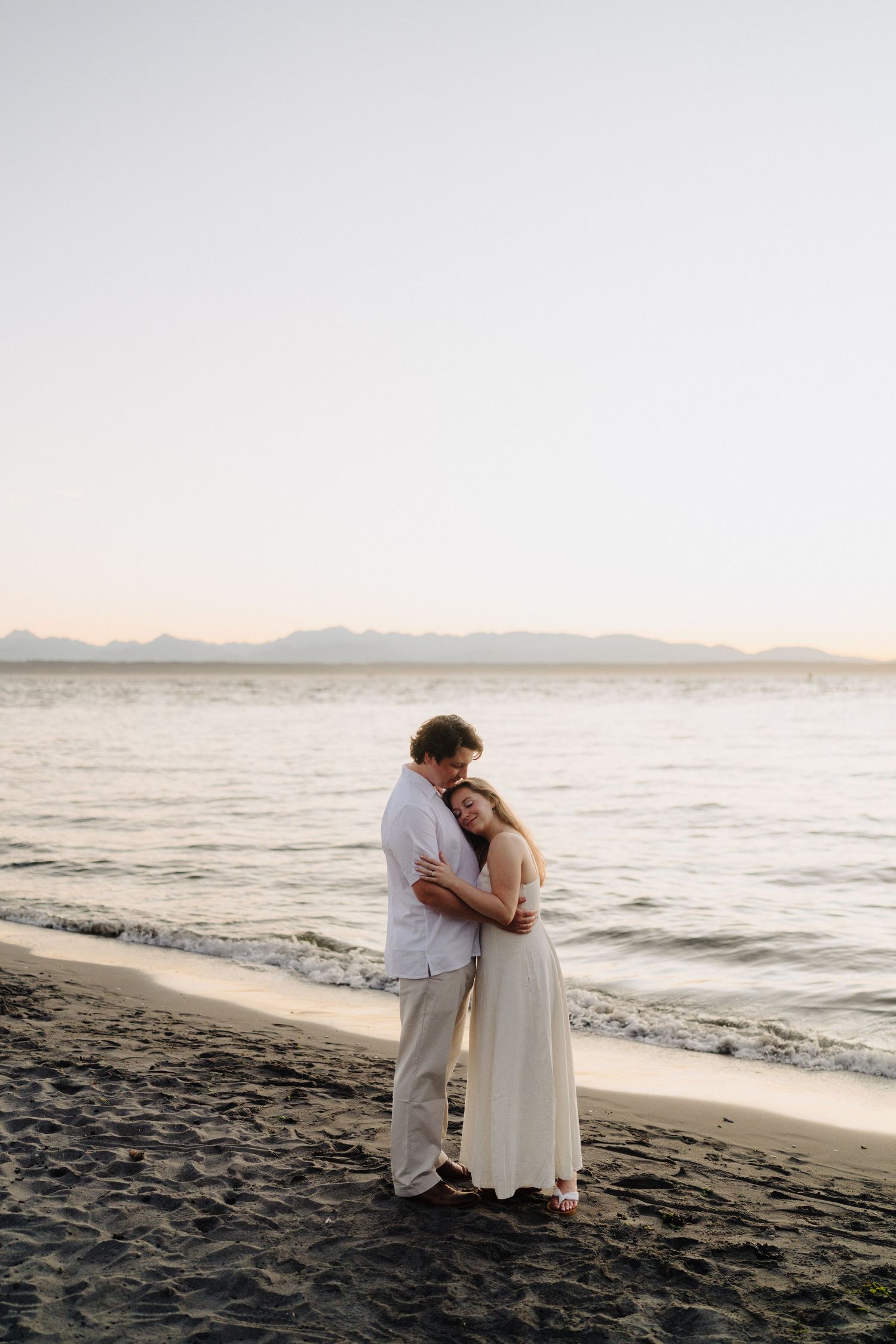 A couple shares a romantic moment on a dark sandy beach with mountains visible in the distance.