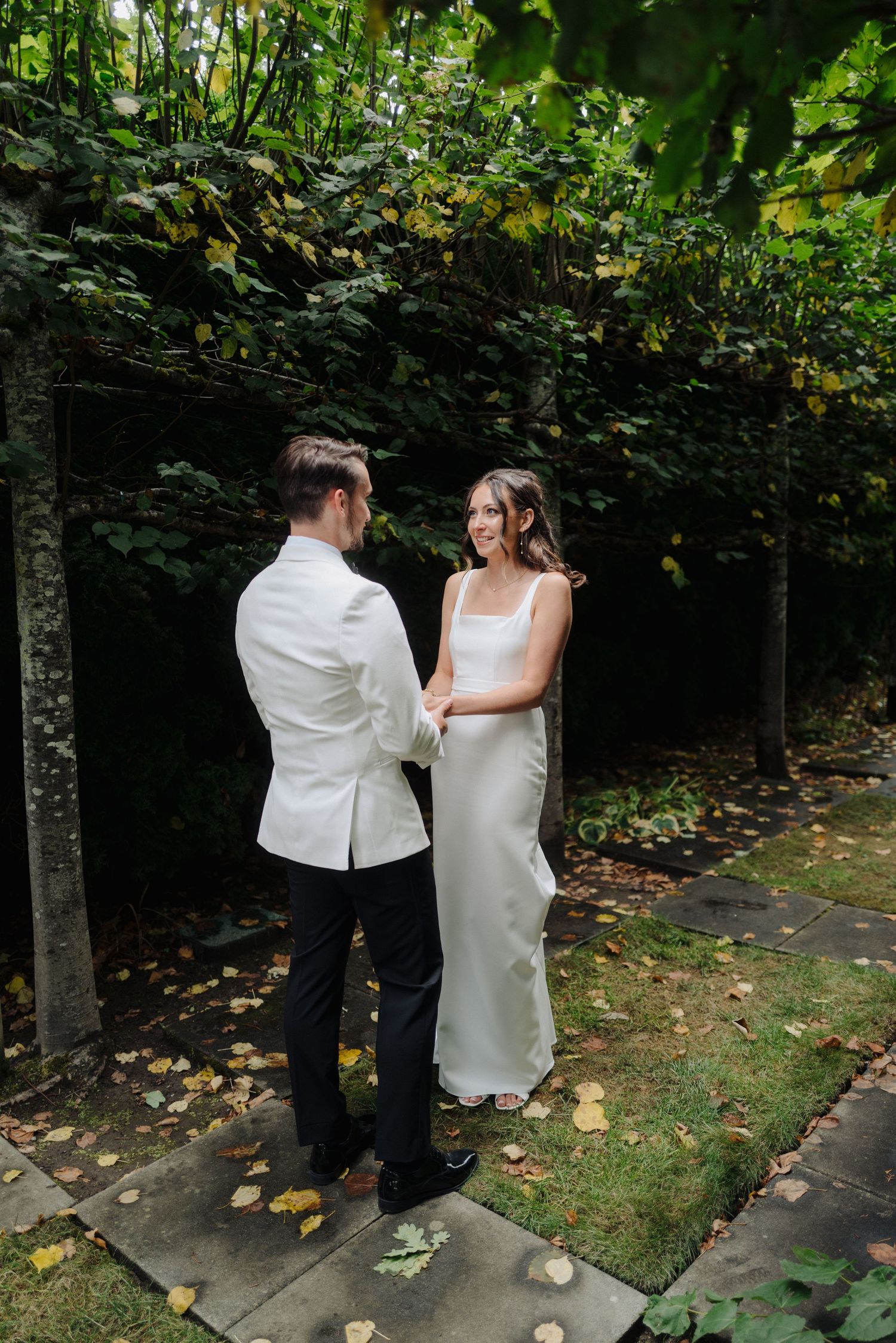 A couple in white formal attire dance together on a garden pathway surrounded by lush greenery.