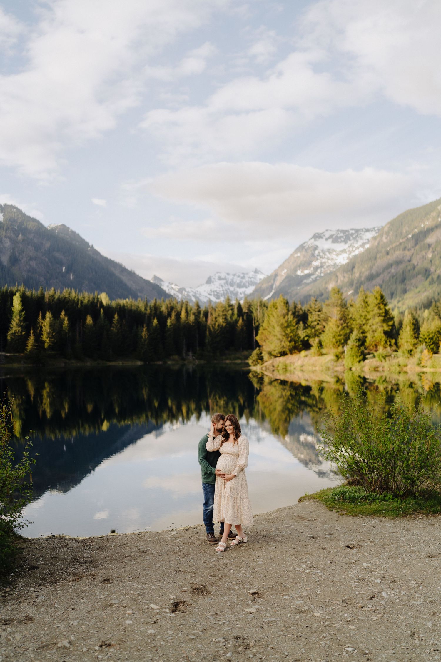 Two people embrace on a rocky lakeshore with majestic mountains and pine trees reflecting in the calm waters.