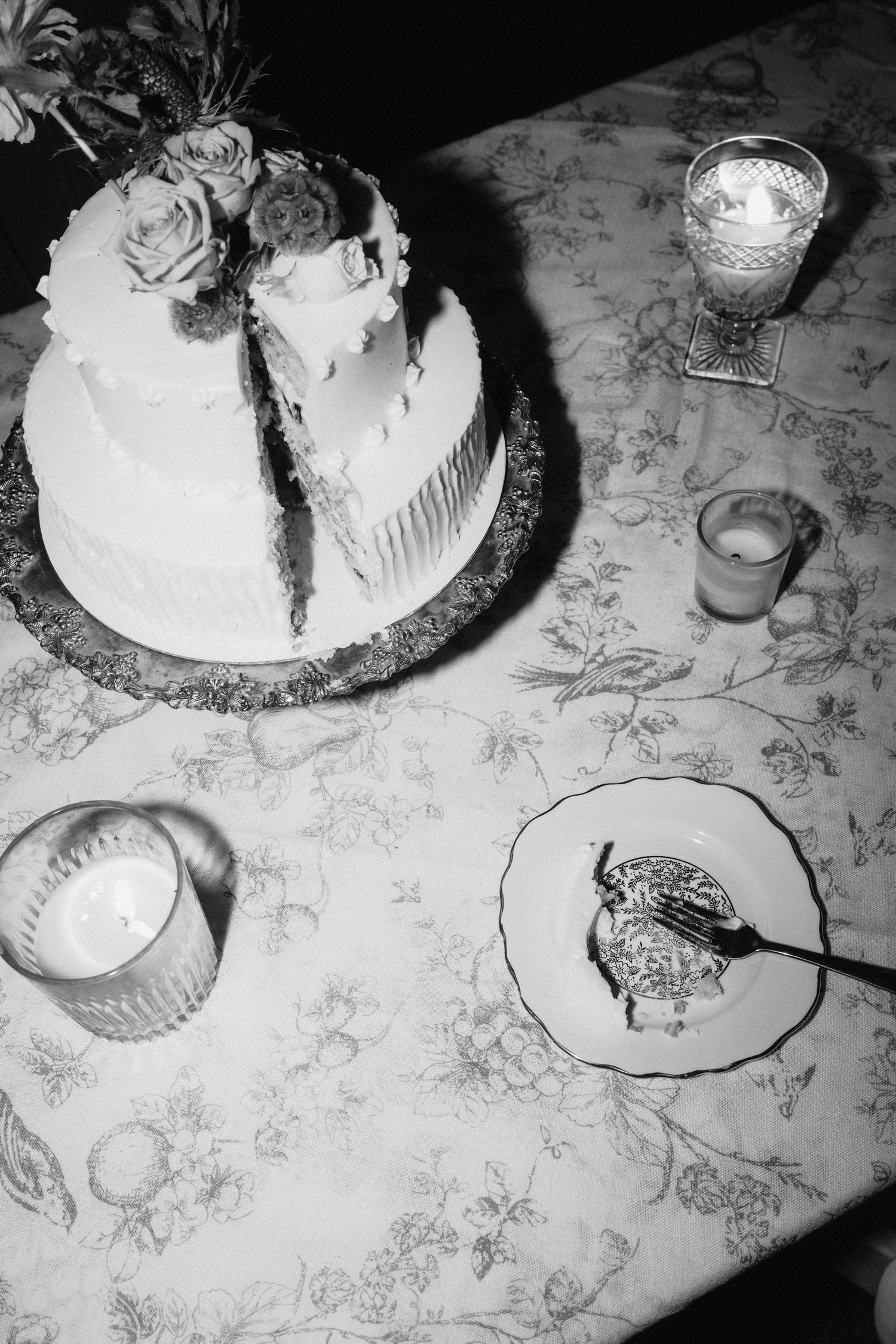 Black and white detail shot of a wedding cake and table settings with floral tablecloth.