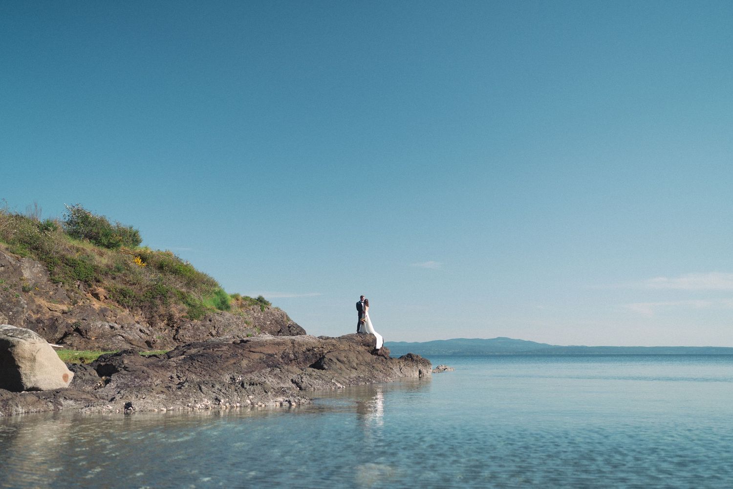 Couple standing on rocky coastal cliffs with calm blue ocean and distant mountains under clear sky.