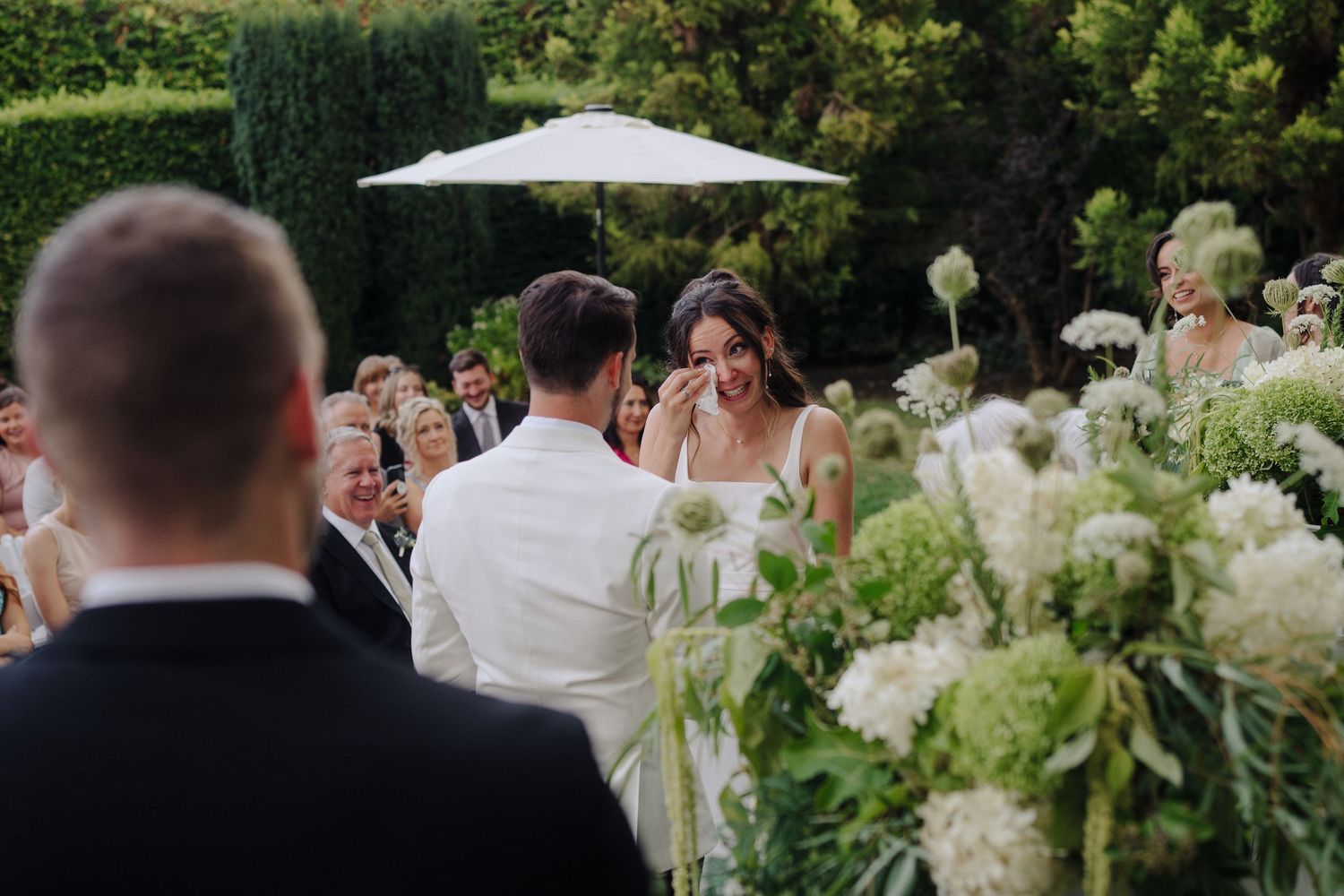 Wedding guests seated at an outdoor ceremony with white floral decorations and umbrellas.
