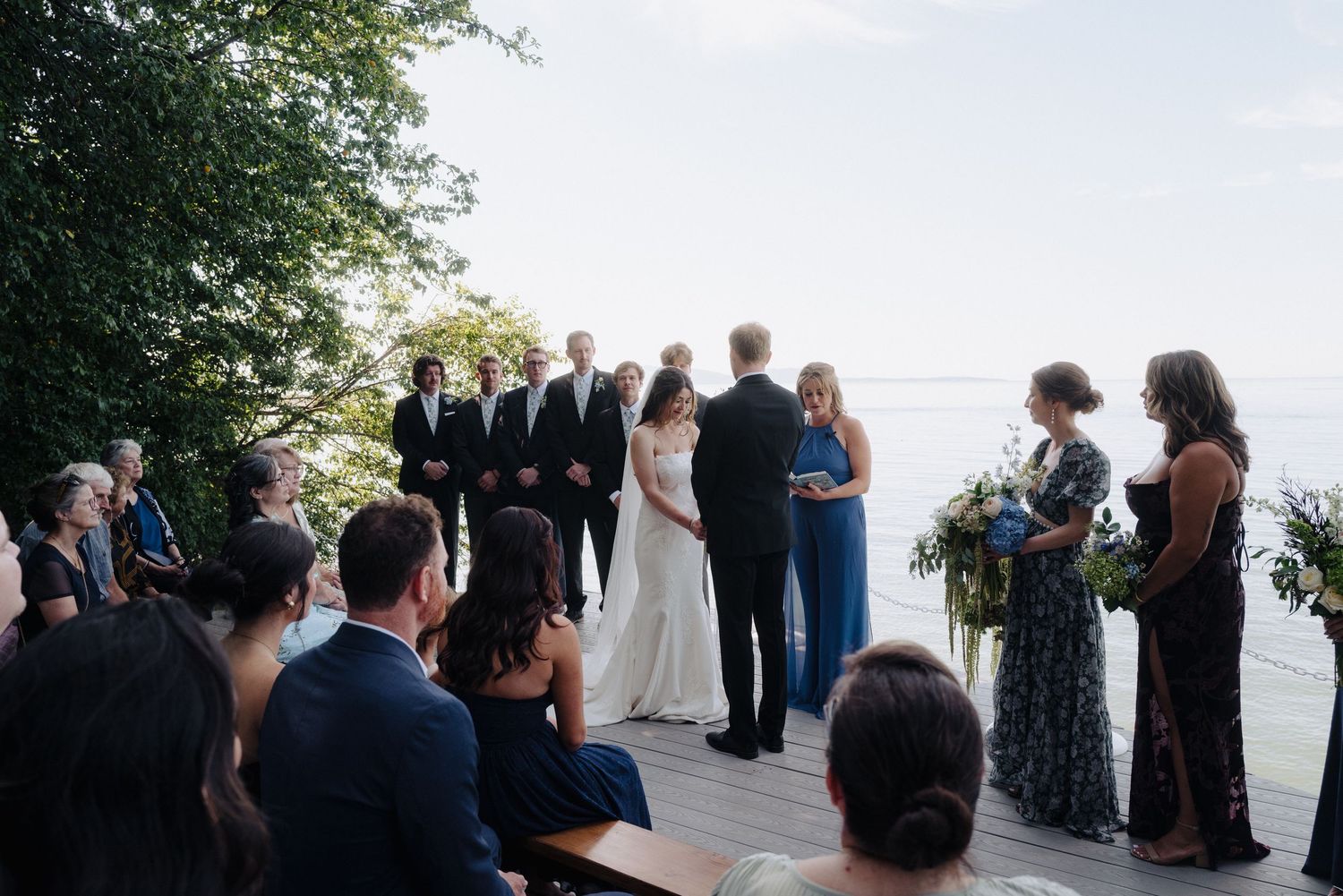 Wedding ceremony guests lined up on a wooden deck with water views and trees in the background.
