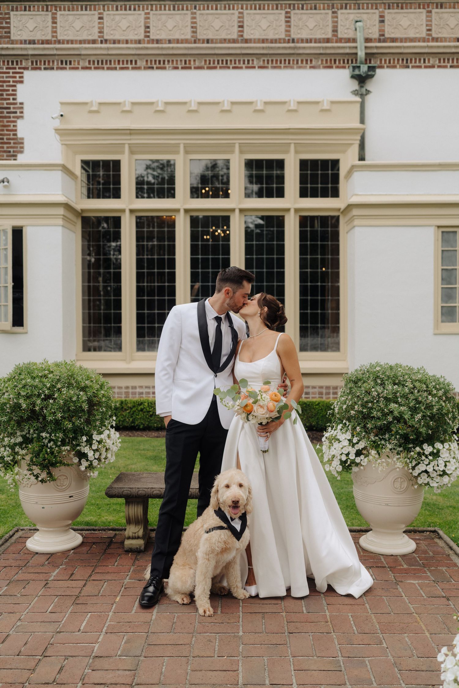 Newlyweds pose with their golden retriever on brick patio outside elegant white mansion.
