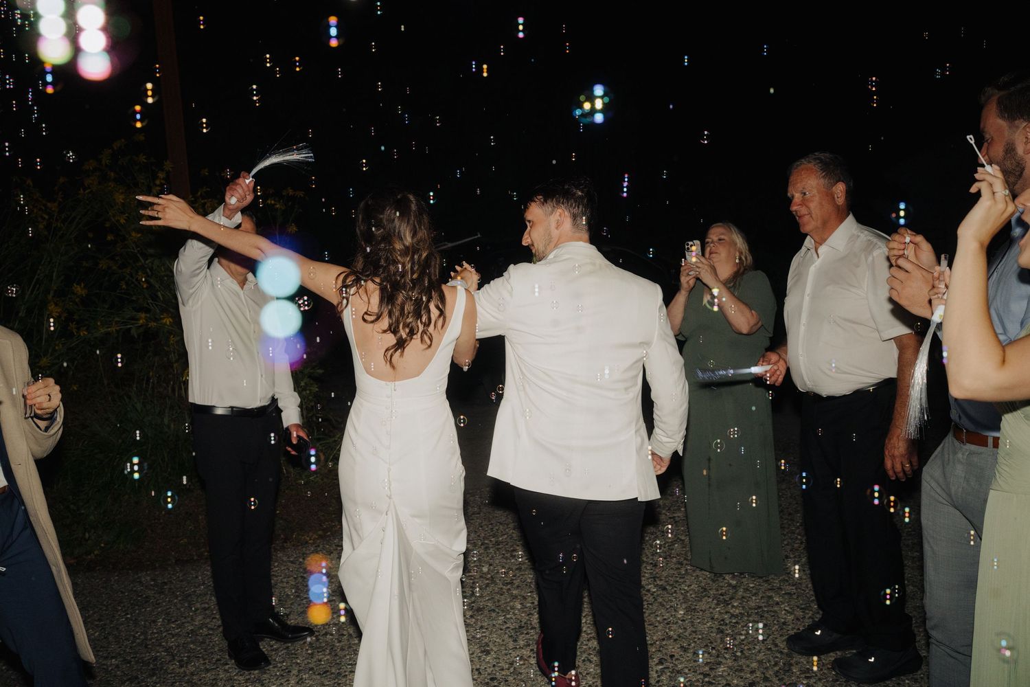 Wedding guests dance and celebrate under twinkling lights at a nighttime outdoor reception.