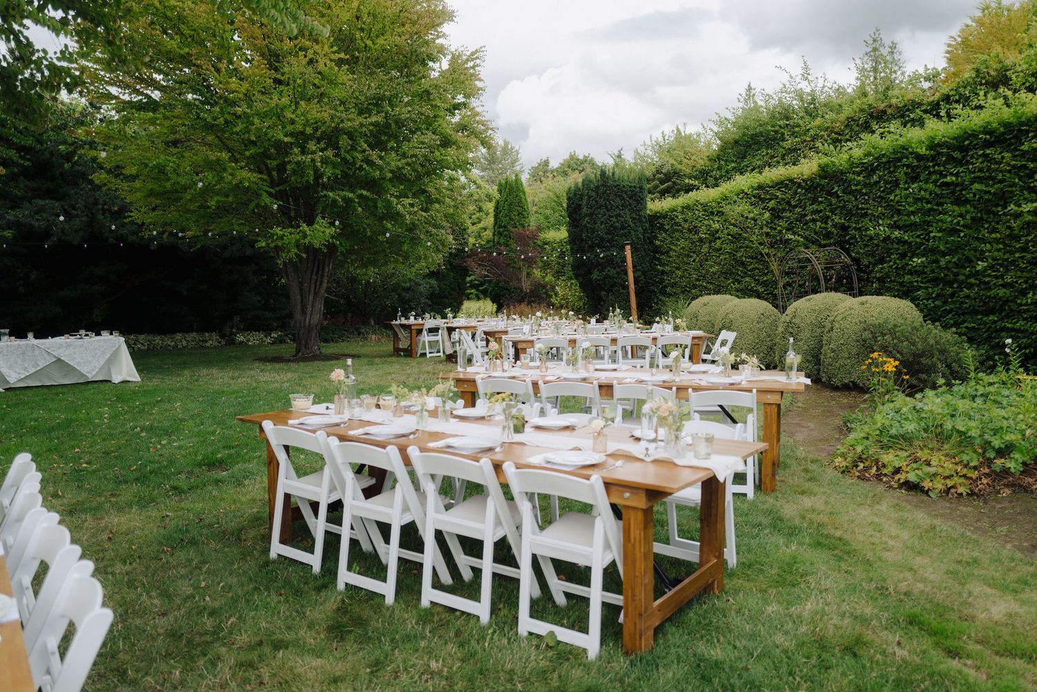 An outdoor wedding reception setup with white chairs and wooden tables in a garden setting.