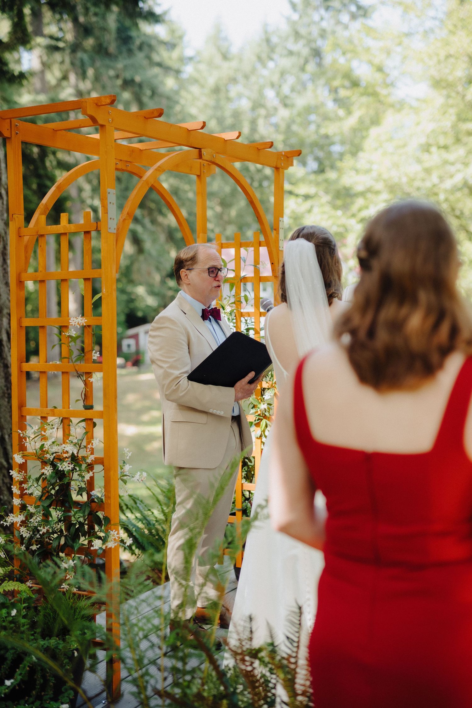 Wedding ceremony under wooden pergola with red-dressed attendees in outdoor garden setting.