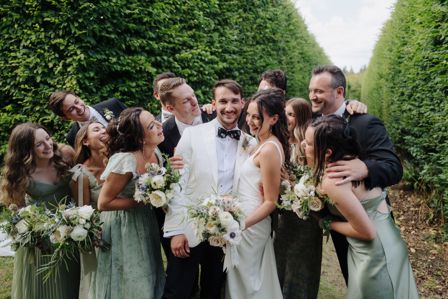 Wedding party poses together in a lush garden pathway with matching attire and floral bouquets.