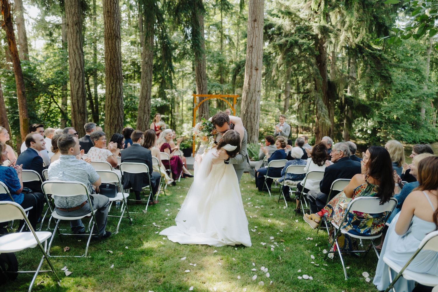 Wedding guests seated in white chairs during an outdoor forest ceremony.