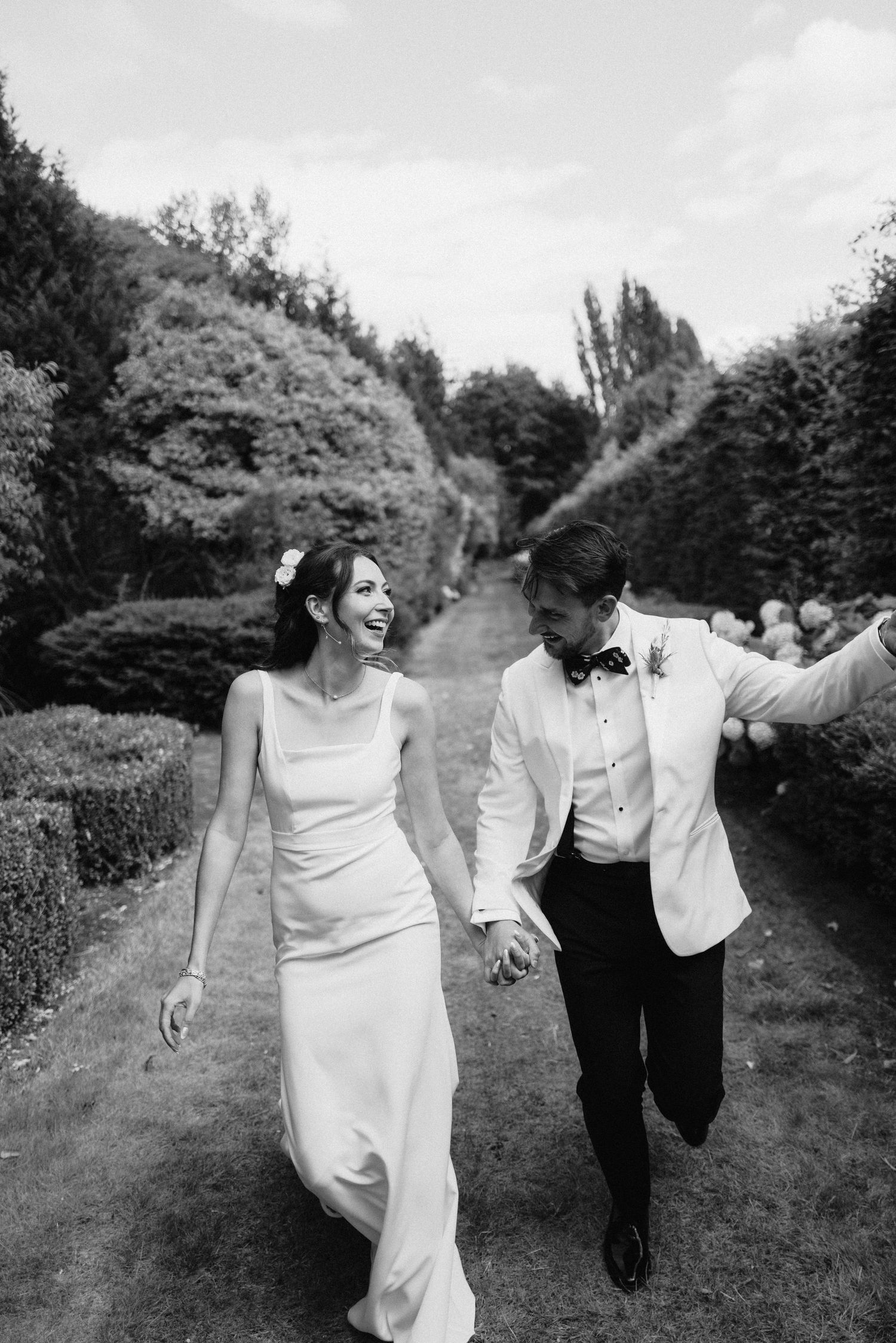 A couple runs playfully together down a tree-lined path in elegant wedding attire.