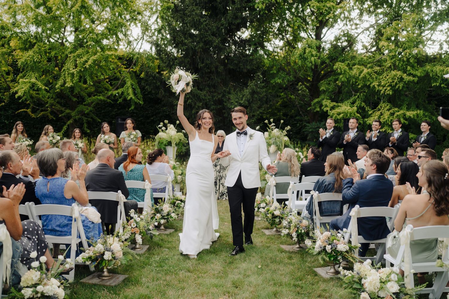 Wide view of wedding guests seated in white chairs during an outdoor garden ceremony.