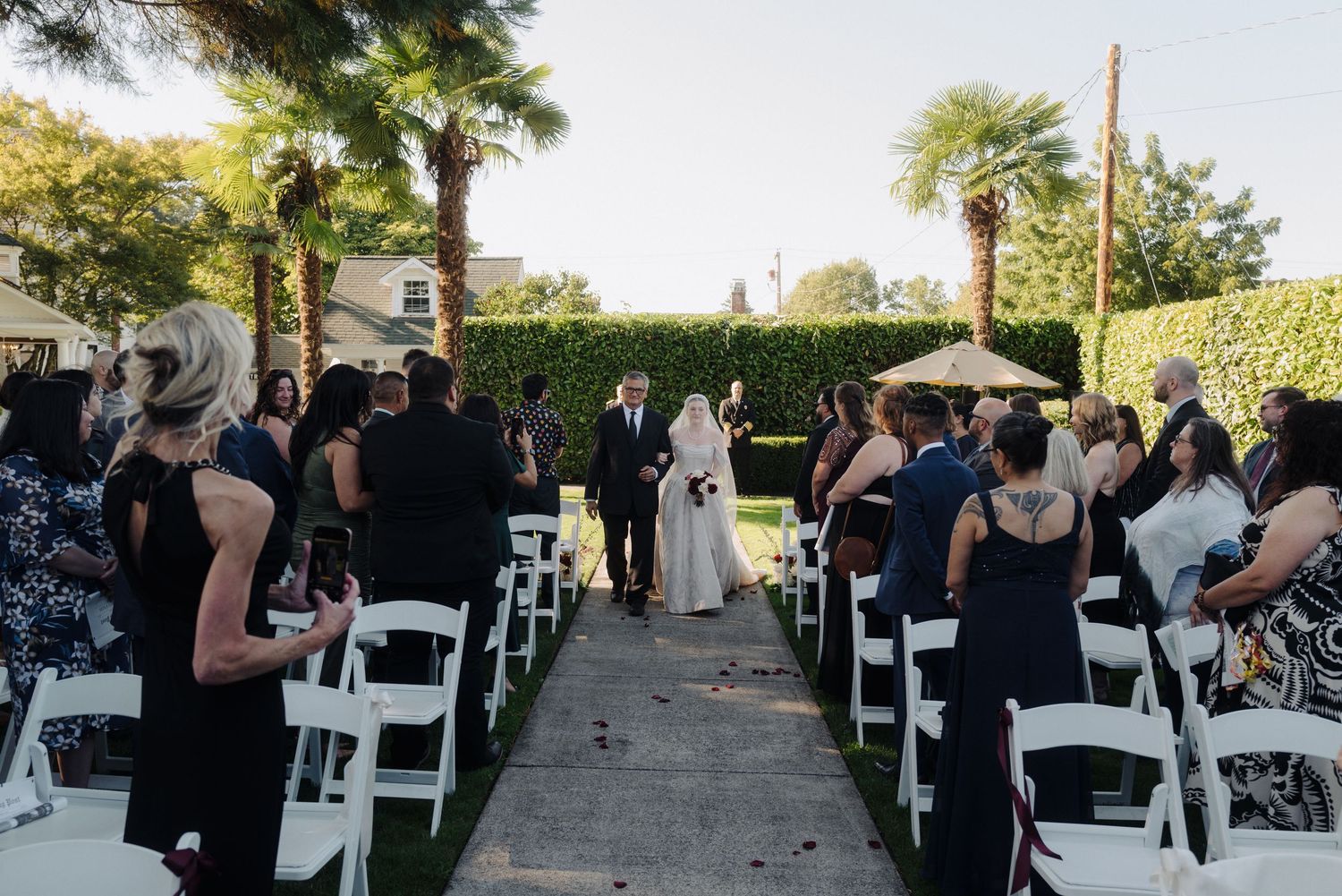Guests seated in white chairs at an outdoor wedding ceremony under trees.