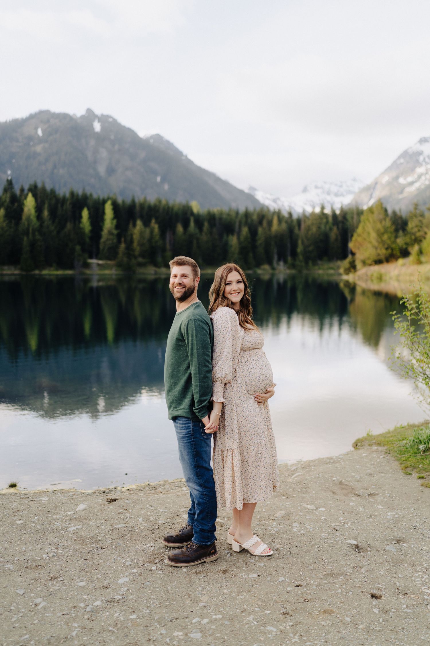 Two people stand together on a rocky shoreline with mountains and a tranquil lake in the background.