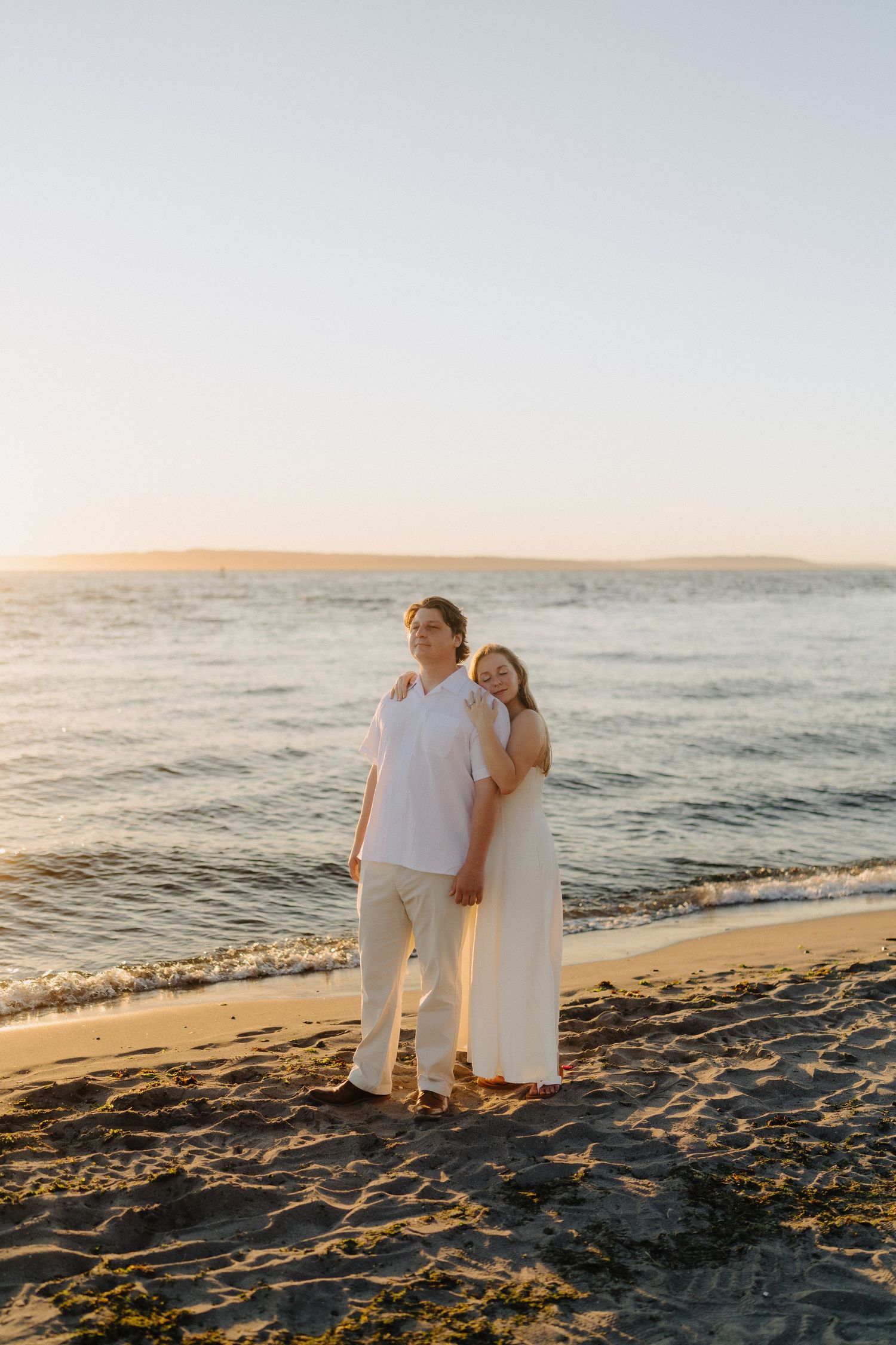 A couple in white outfits embracing on a sandy beach during golden hour sunset.