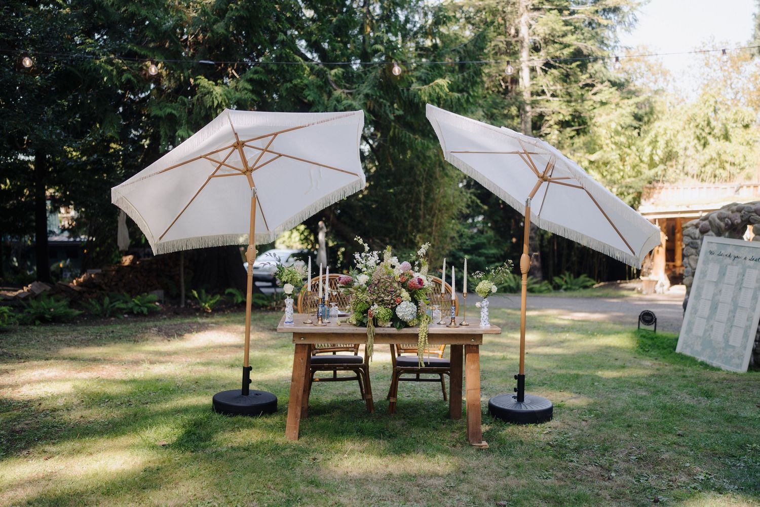 Outdoor reception setup with wooden table and white umbrellas in a sunny garden setting.