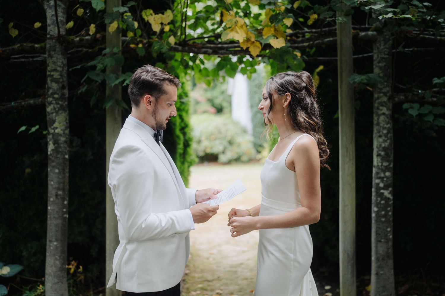 A couple walks together along a tree-lined path in formal wedding attire.