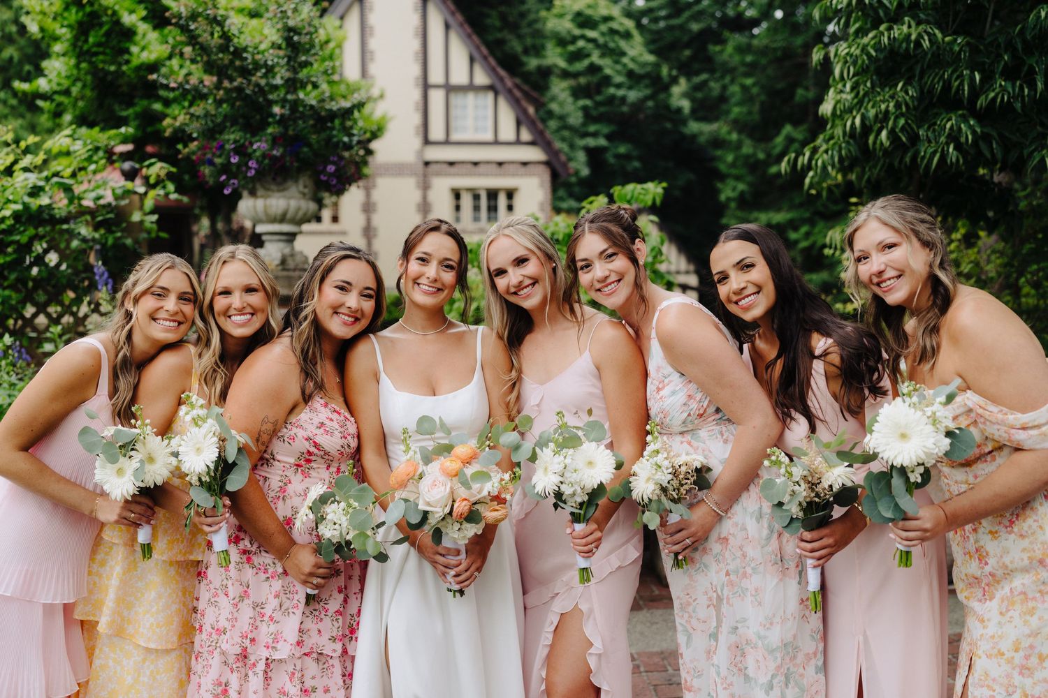 Bridesmaids in pastel dresses holding floral bouquets pose together outdoors.