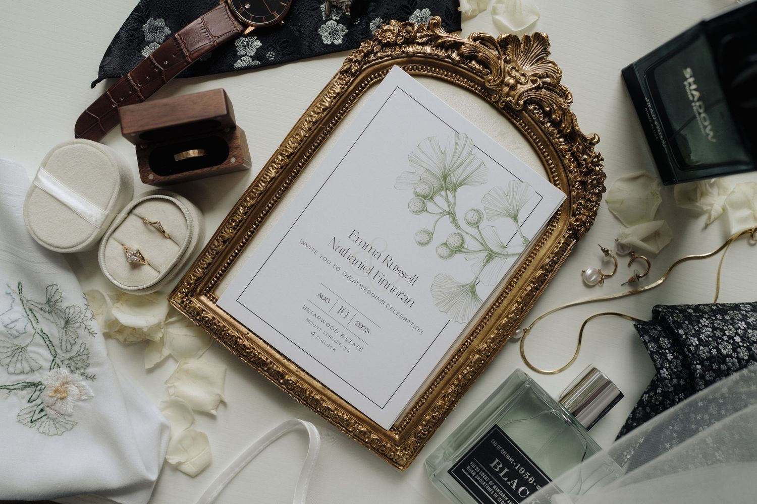 Vintage gold frame displaying wedding invitation surrounded by perfume bottles and accessories.