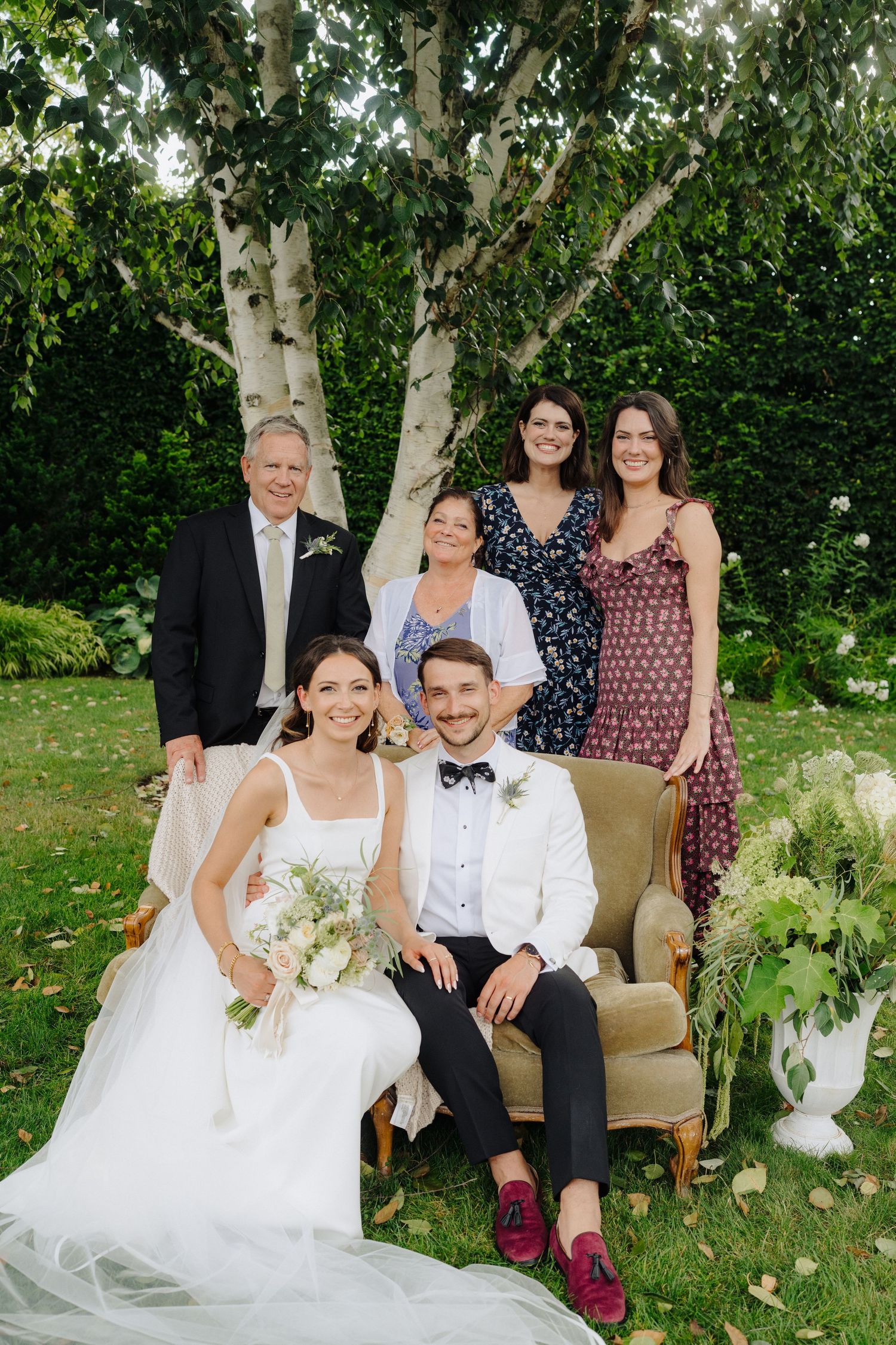 Wedding party poses together on vintage furniture outdoors with birch trees and greenery in background.