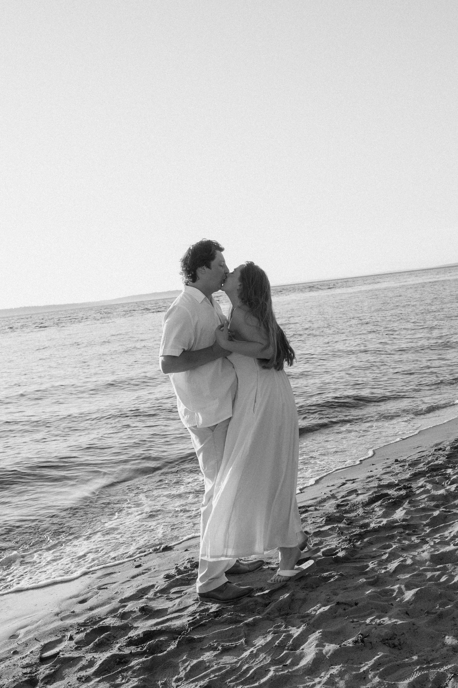 Two people in white clothing share an intimate moment on a rocky beach at sunset.