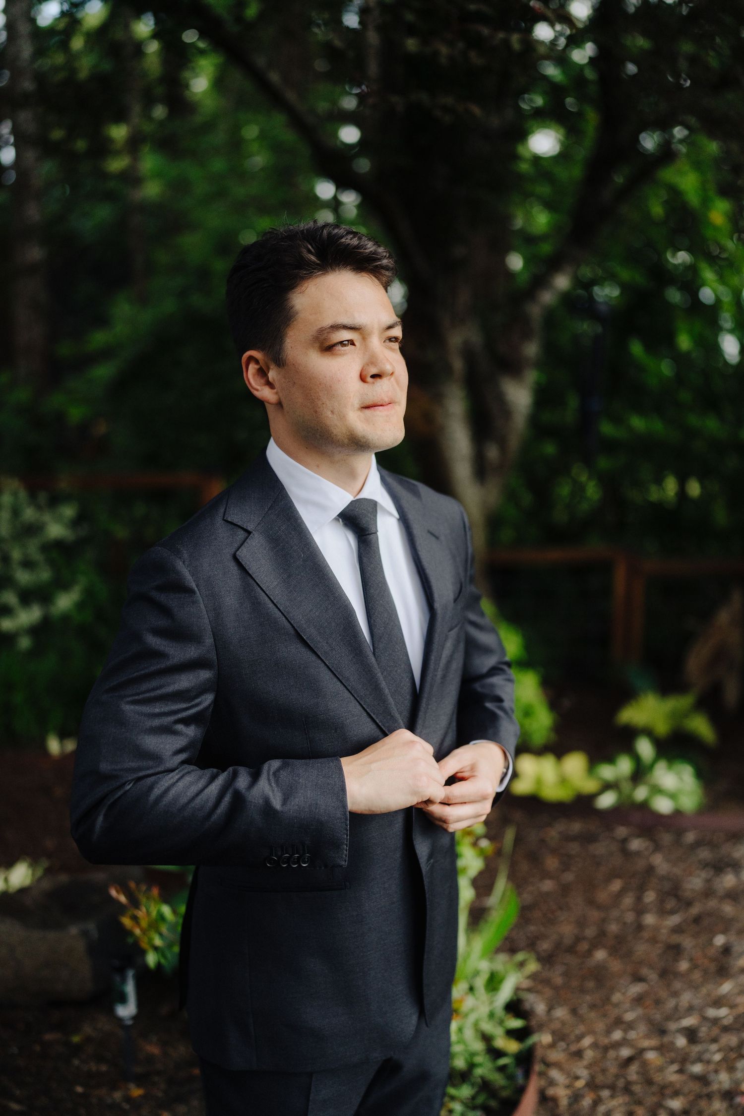 A groom in a black suit and tie adjusts his jacket while standing outdoors in a garden setting.