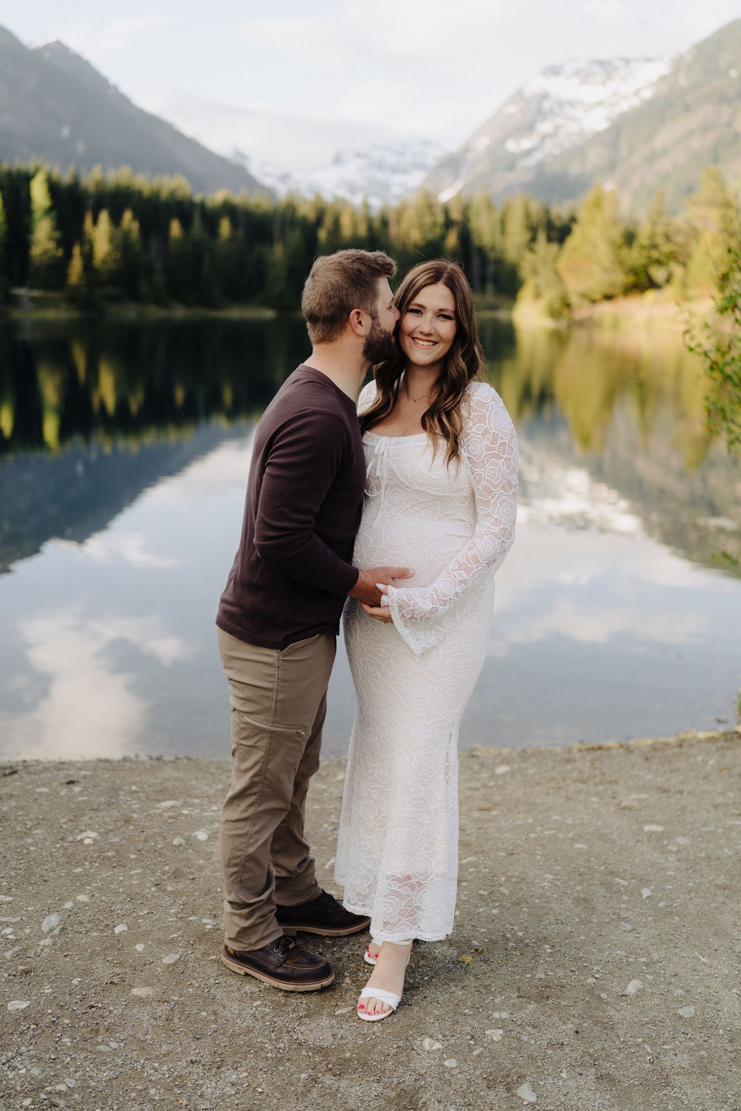 A romantic lakeside scene with a couple embracing on a rocky shore with mountains and forest in the background.