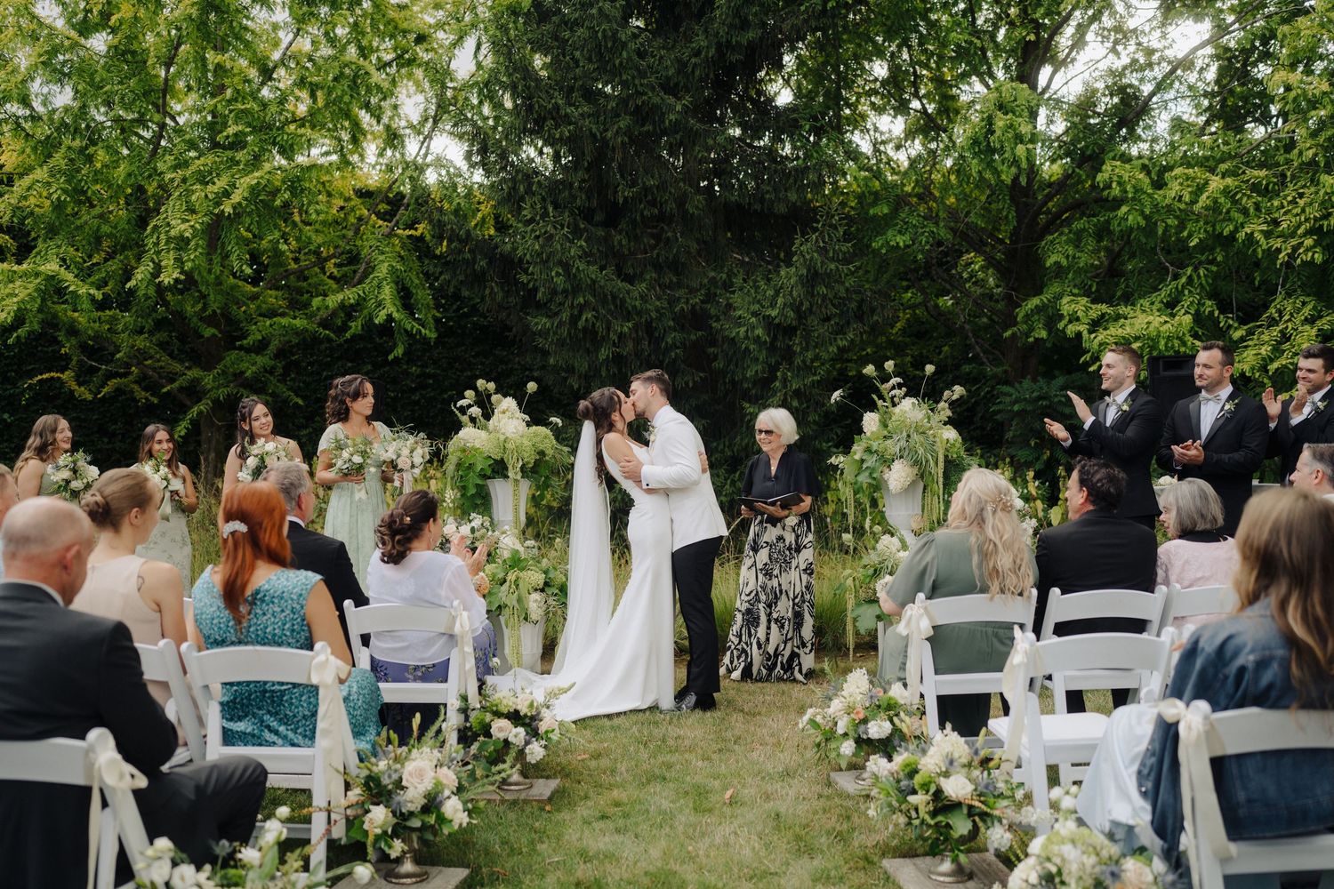 Outdoor wedding ceremony with white chairs and natural greenery backdrop.