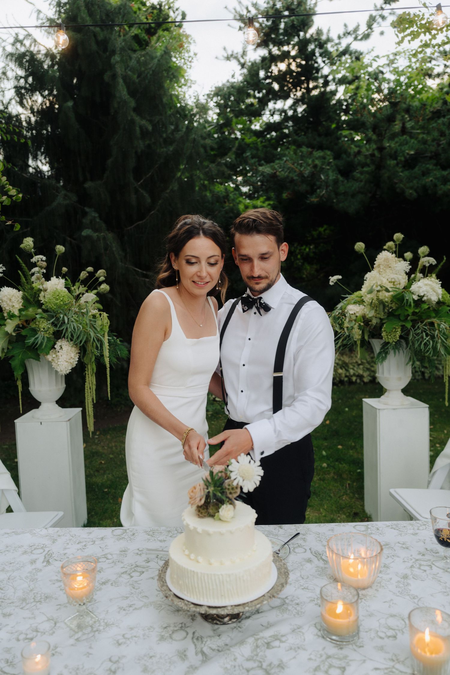 A couple cuts their wedding cake at an elegant outdoor reception with white floral arrangements.