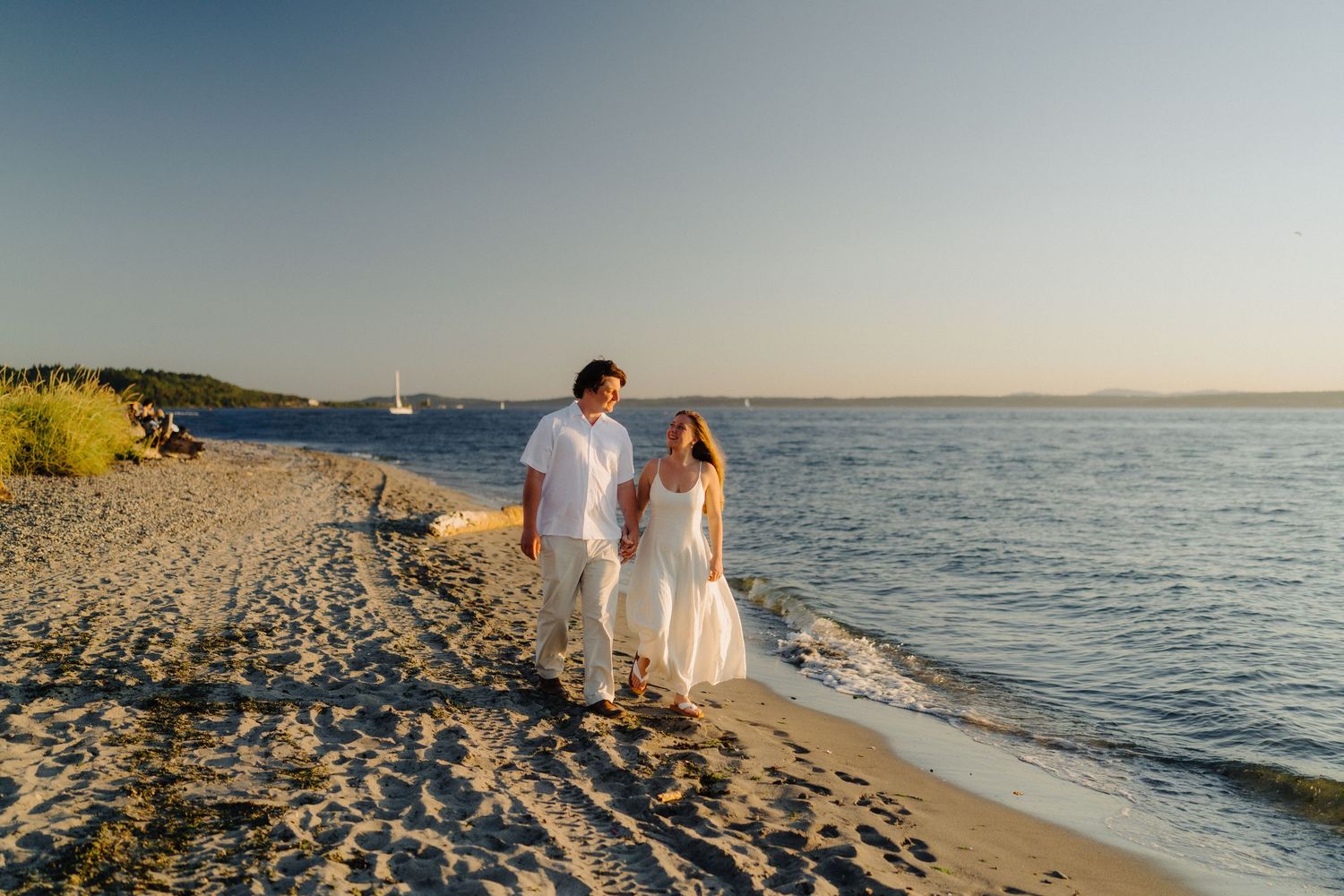 Romantic couple in white outfits walking along a scenic beach at sunset with ocean views.