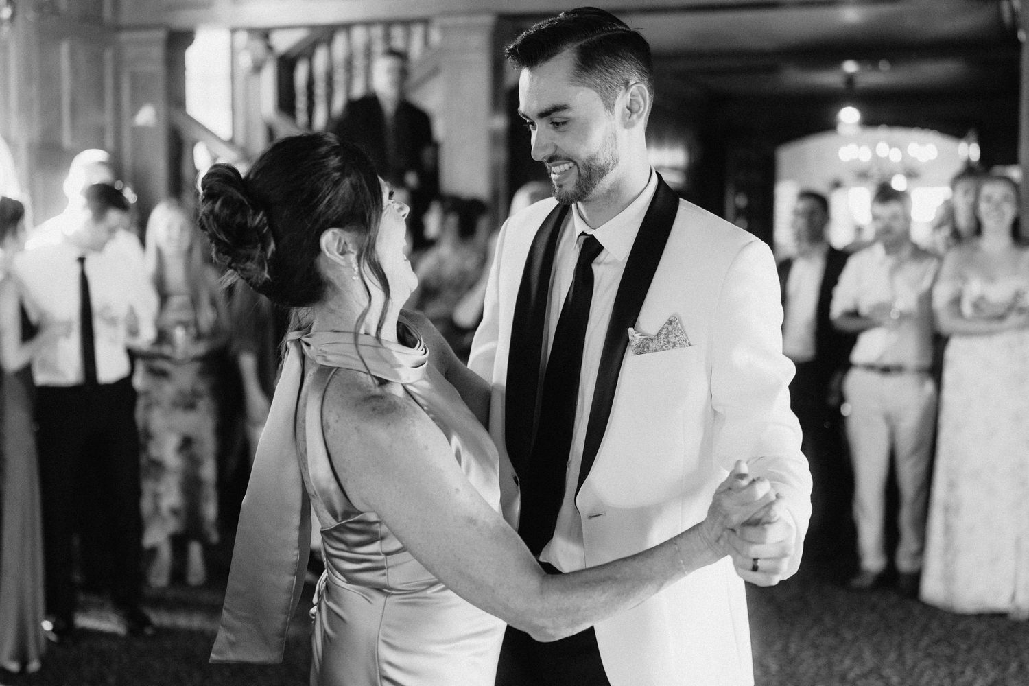 Wedding couple sharing first dance surrounded by guests in dimly lit reception venue.
