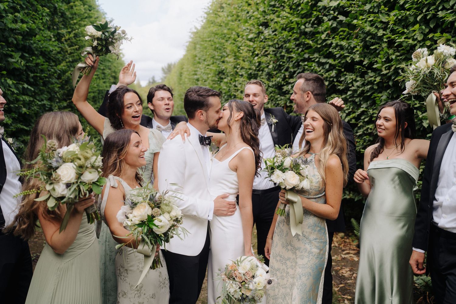 Wedding party members in formal attire laugh together while holding white floral bouquets outdoors.