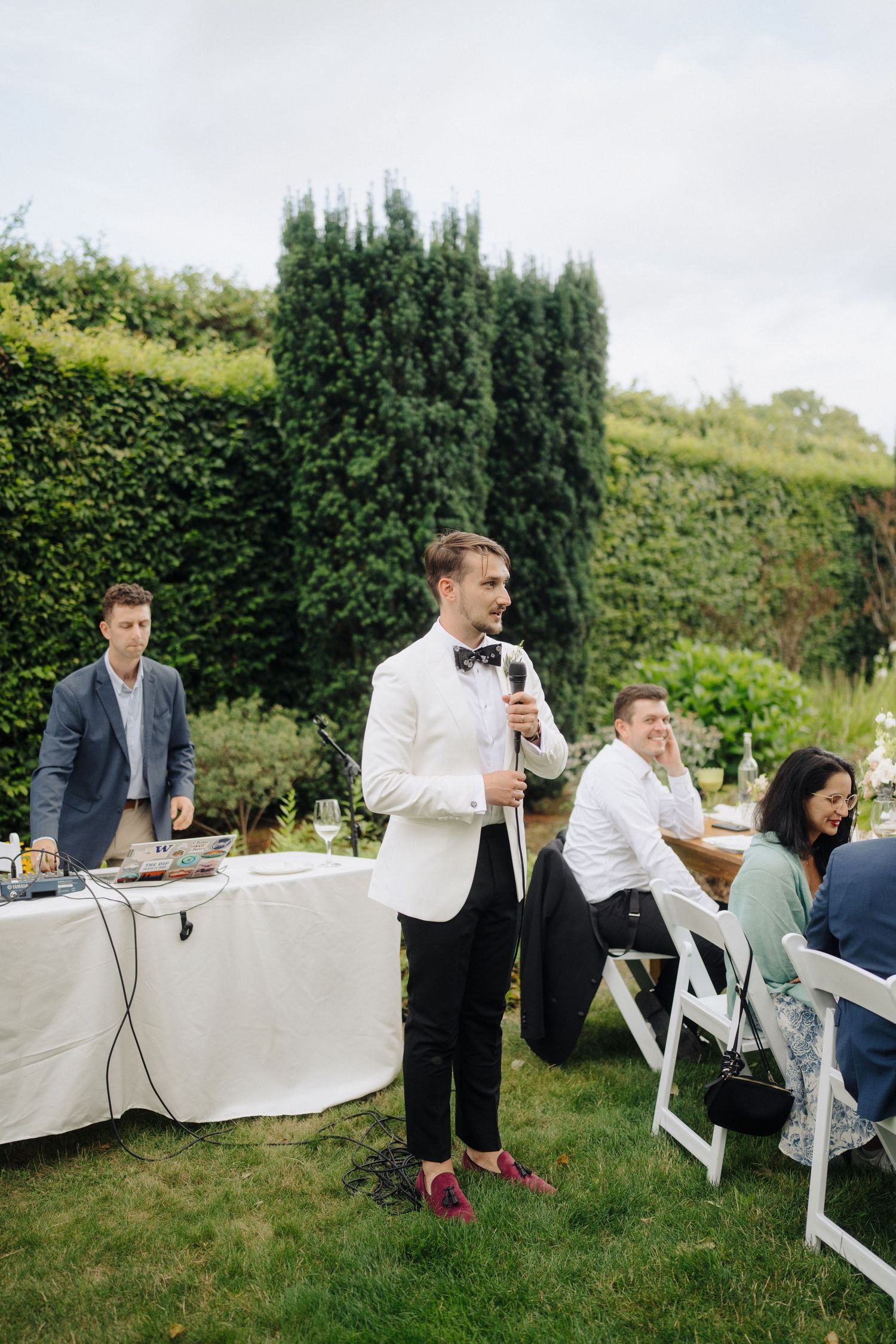 A wedding speech being given outdoors with a hedge backdrop and white-clothed tables at a garden reception.