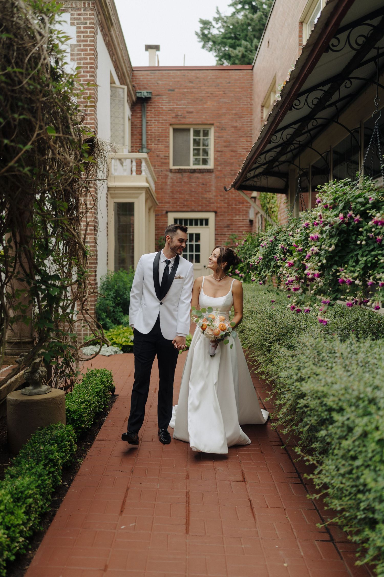 Wedding couple strolling along a red brick walkway surrounded by garden landscaping.
