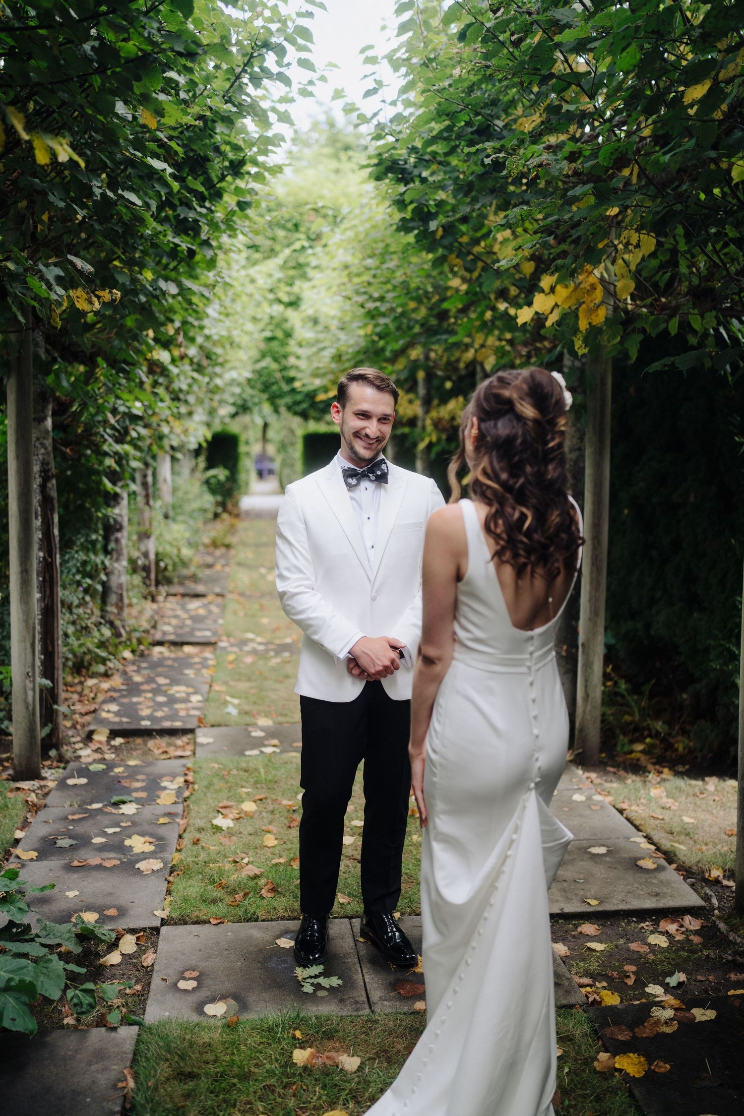 A couple in wedding attire shares an intimate moment along a tree-lined path covered in fallen leaves.