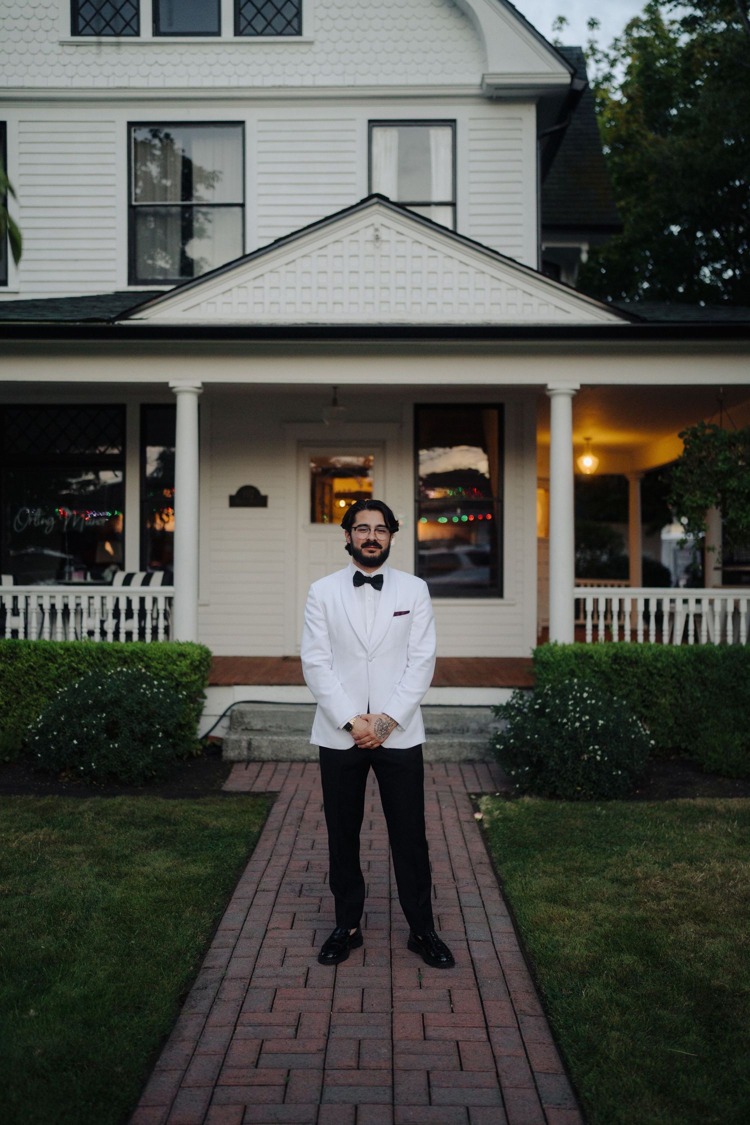 A person in a white tuxedo jacket poses on a brick walkway in front of a traditional white house.