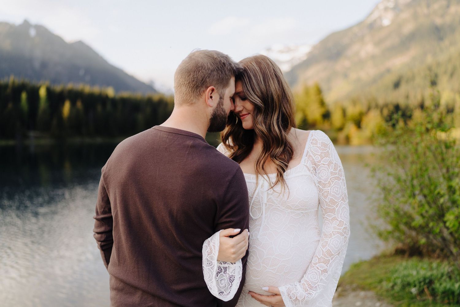 Couple in white and brown outfits posing by a mountain lake during golden hour.