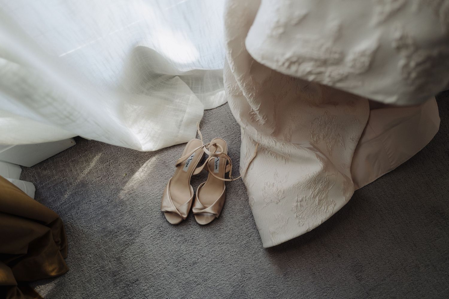 A pair of elegant beige sandals rests on a dark floor next to flowing white fabric.